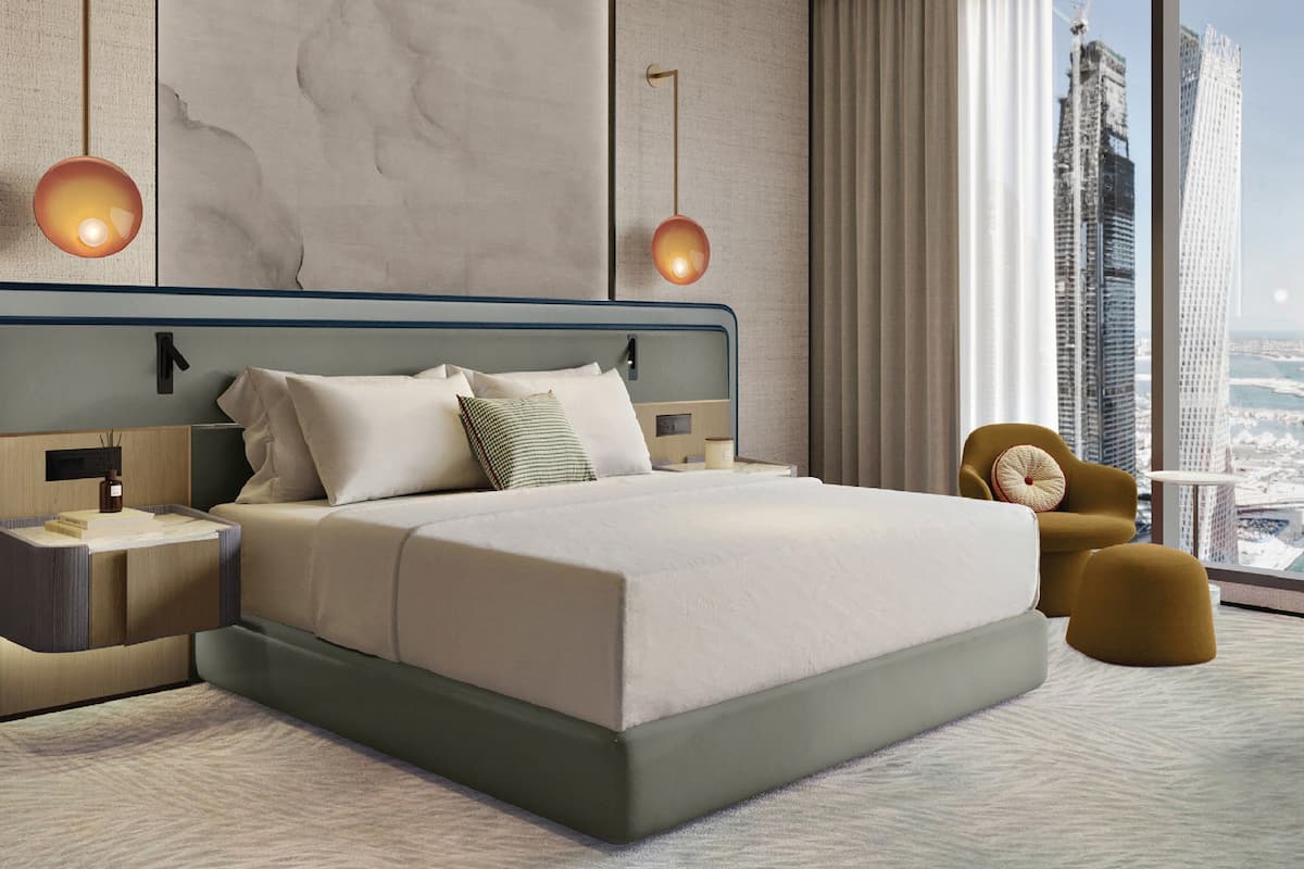 A modern hotel room with a large bed, two bedside lamps, a nightstand, a chair, and a view of the cityscape.