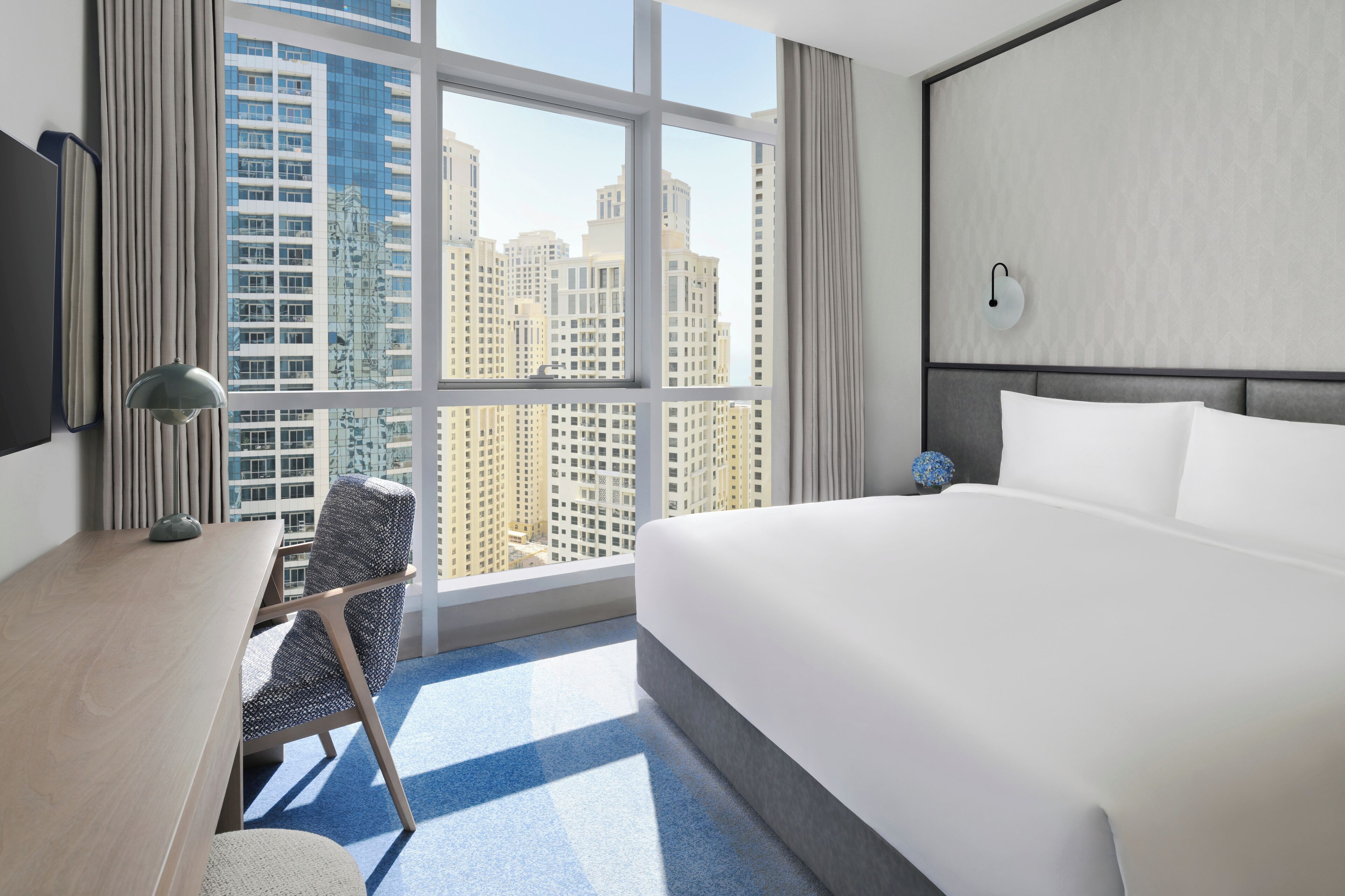 A hotel room with a large bed, a desk with a chair, a TV, and a window with a city view.