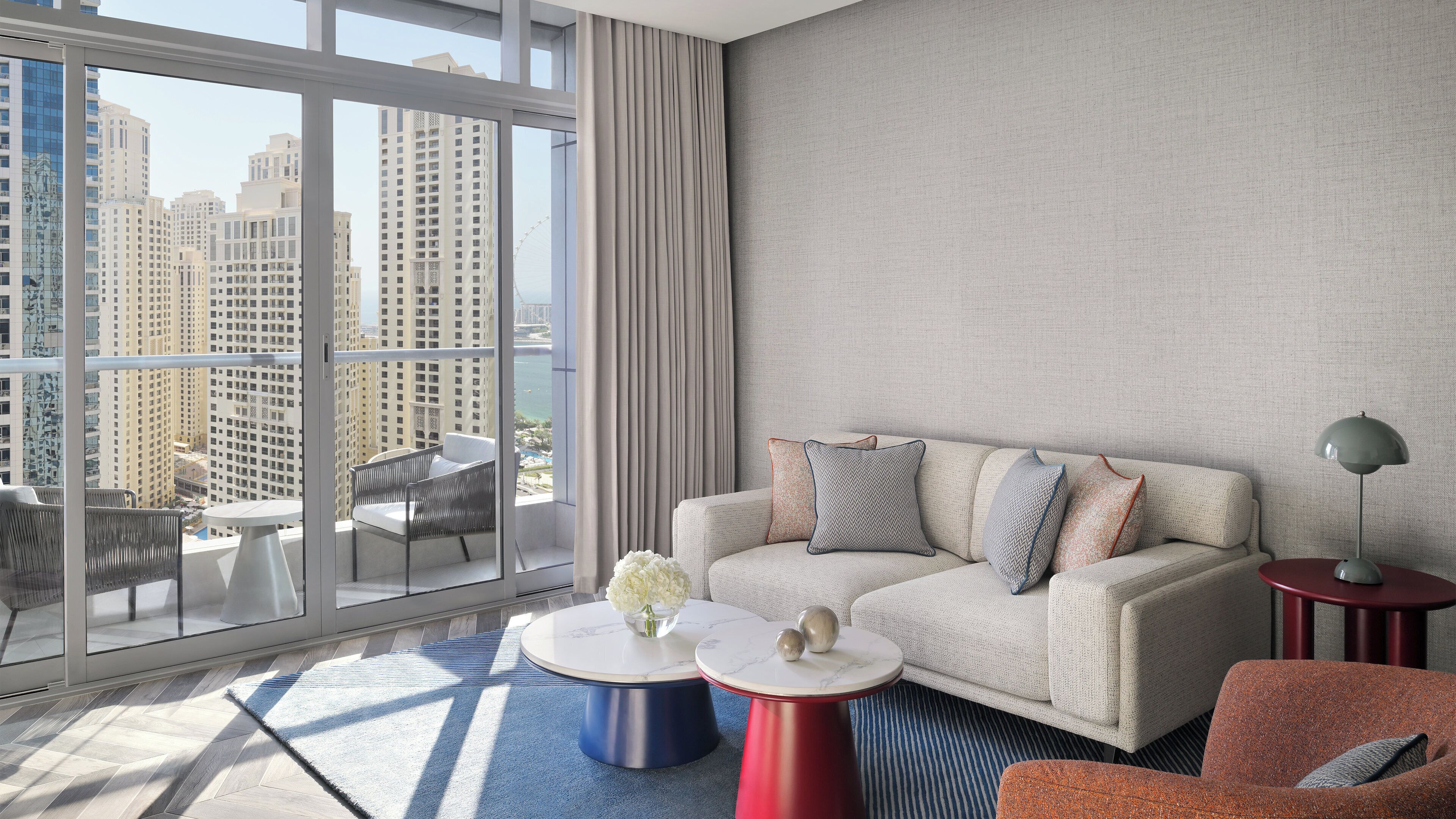 A modern hotel room with a balcony, a sofa set, two round tables, and a view of high-rise buildings.