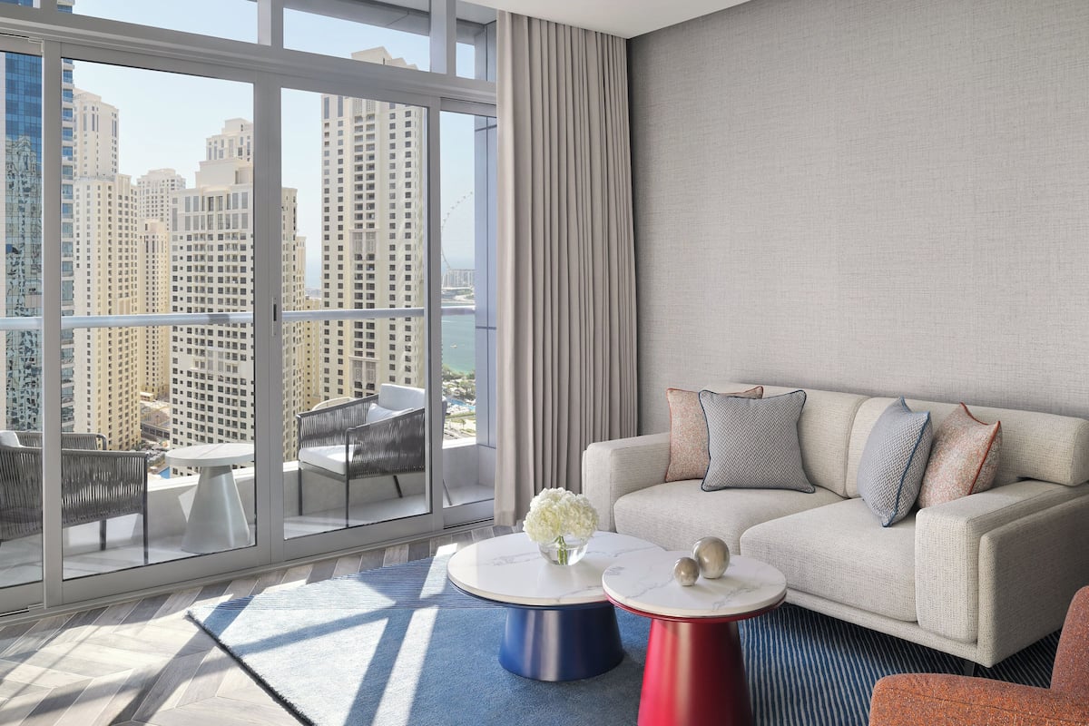 A modern hotel room with a balcony, a sofa set, two round tables, and a view of high-rise buildings.