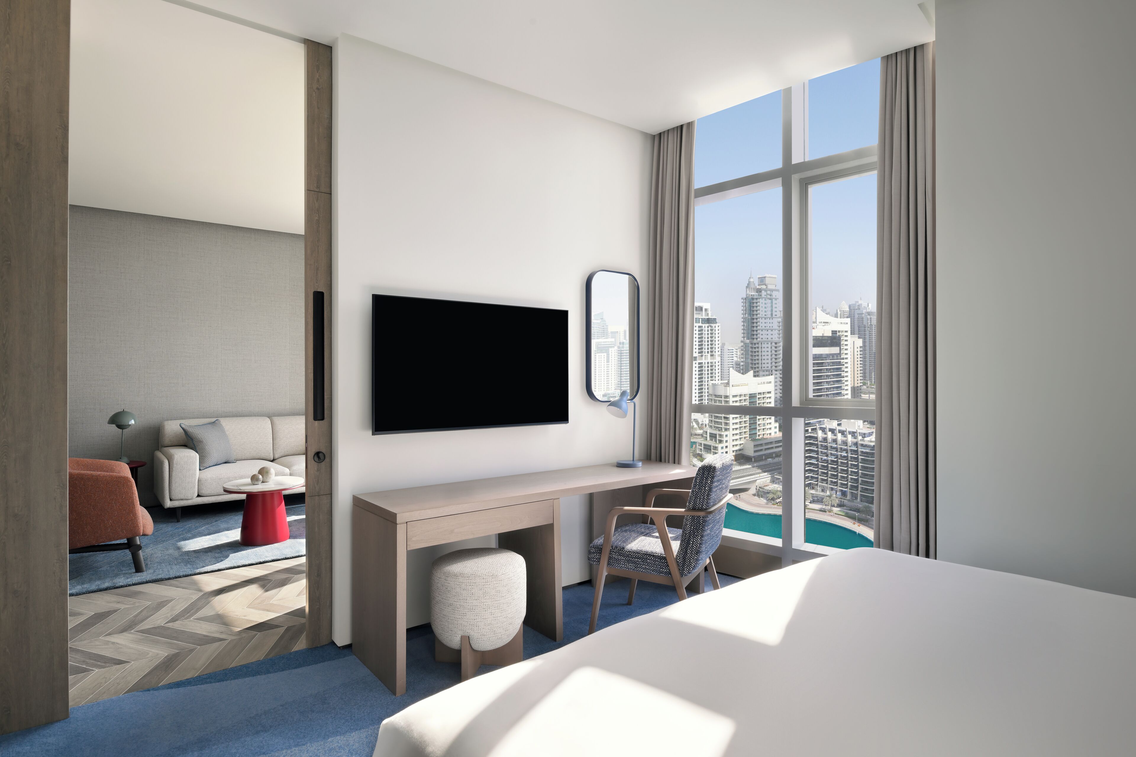 A modern hotel room with a large bed, a desk with a chair, a TV, and a view of the cityscape through large windows.