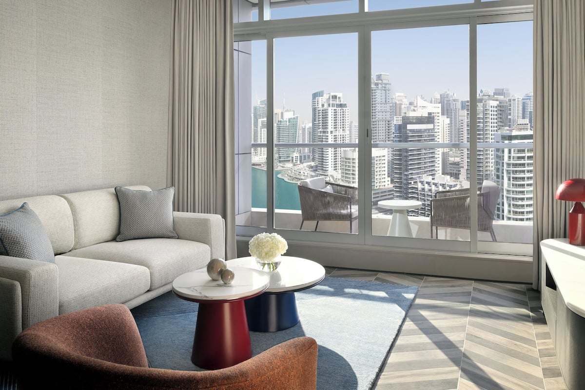 A modern living room with a sofa, a round coffee table, a red lamp, and a view of a cityscape through large windows.