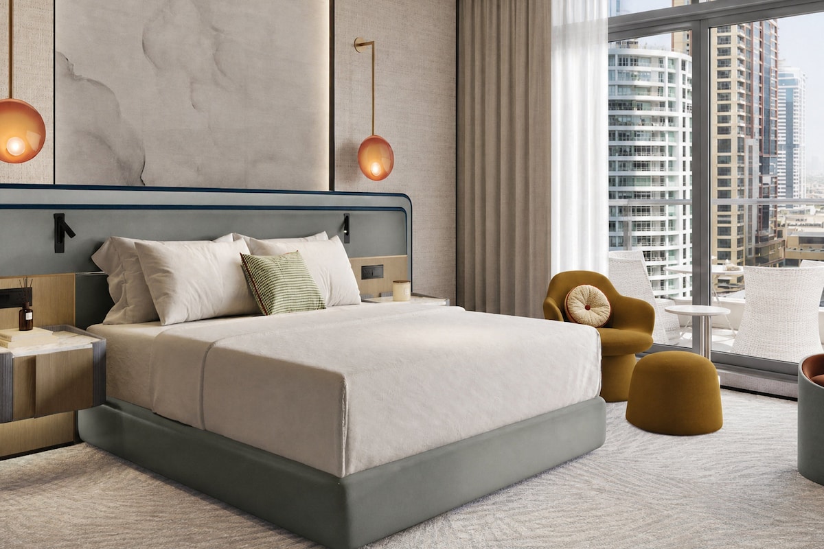 Quarto de hotel moderno com uma cama grande, uma mesa de cabeceira, um espaço de estar com cadeiras e vista para a cidade através de grandes janelas.