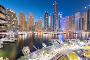 A waterfront cityscape with high-rise buildings, a marina with yachts, and a well-lit promenade.