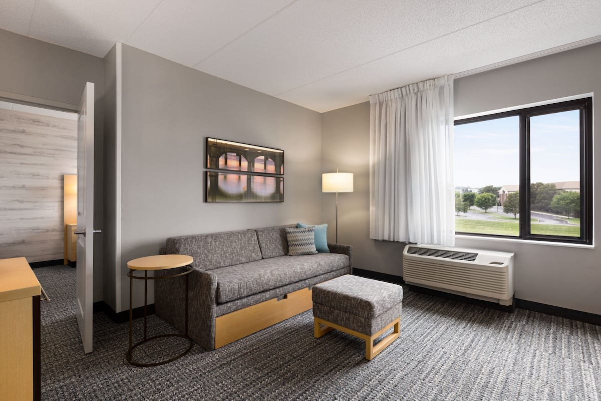 Una habitación de hotel moderna con un sofá gris, una pequeña mesa redonda, una mesita auxiliar, una lámpara, una ventana con vistas y un equipo de aire acondicionado.
