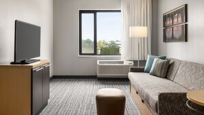 Une chambre d’hôtel avec un téléviseur à écran plat posé sur un meuble en bois, un canapé gris agrémenté de coussins à motifs, une petite table ronde et une fenêtre donnant sur des arbres et des immeubles.