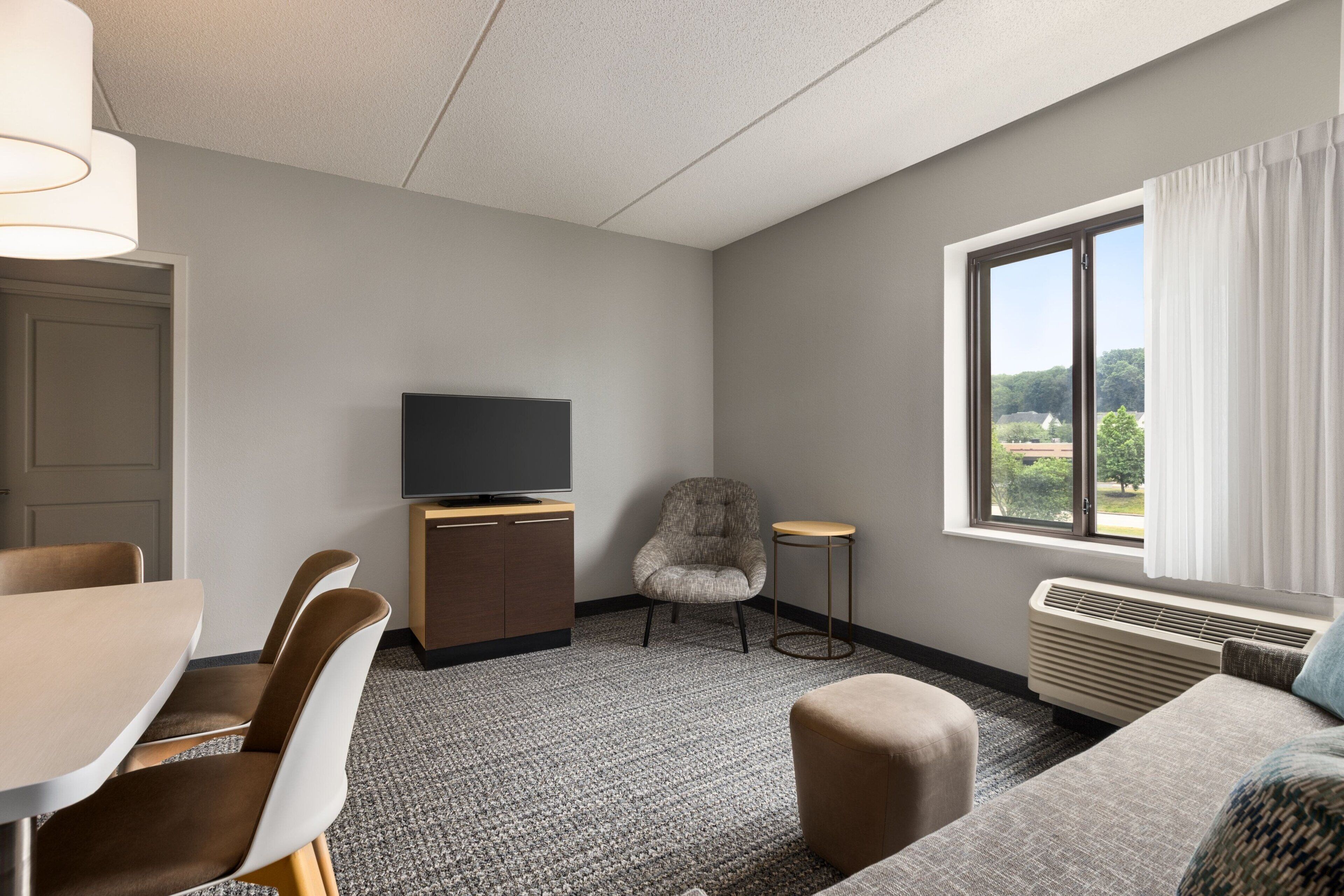A hotel room with a dining area, a television, a chair, a window with a view, and a heater.