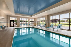 Une piscine intérieure à l’eau d’un bleu cristallin, entourée de chaises longues et dotée de grandes fenêtres offrant une vue sur l’extérieur.