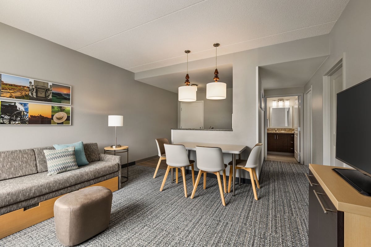 Una habitación de hotel moderna con alfombra gris, un sofá gris con cojines azul y blanco, una mesa de centro de madera, un reposapiés gris, una zona de comedor con mesa y sillas blancas, un espejo grande sobre la mesa y un televisor de pantalla plana sobre un mueble de madera.