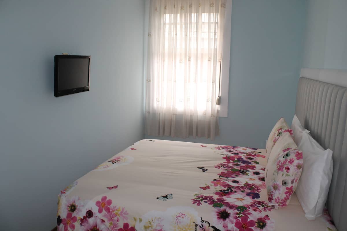 Een slaapkamer met een bed, een aan de muur bevestigde televisie en een raam met dunne gordijnen.