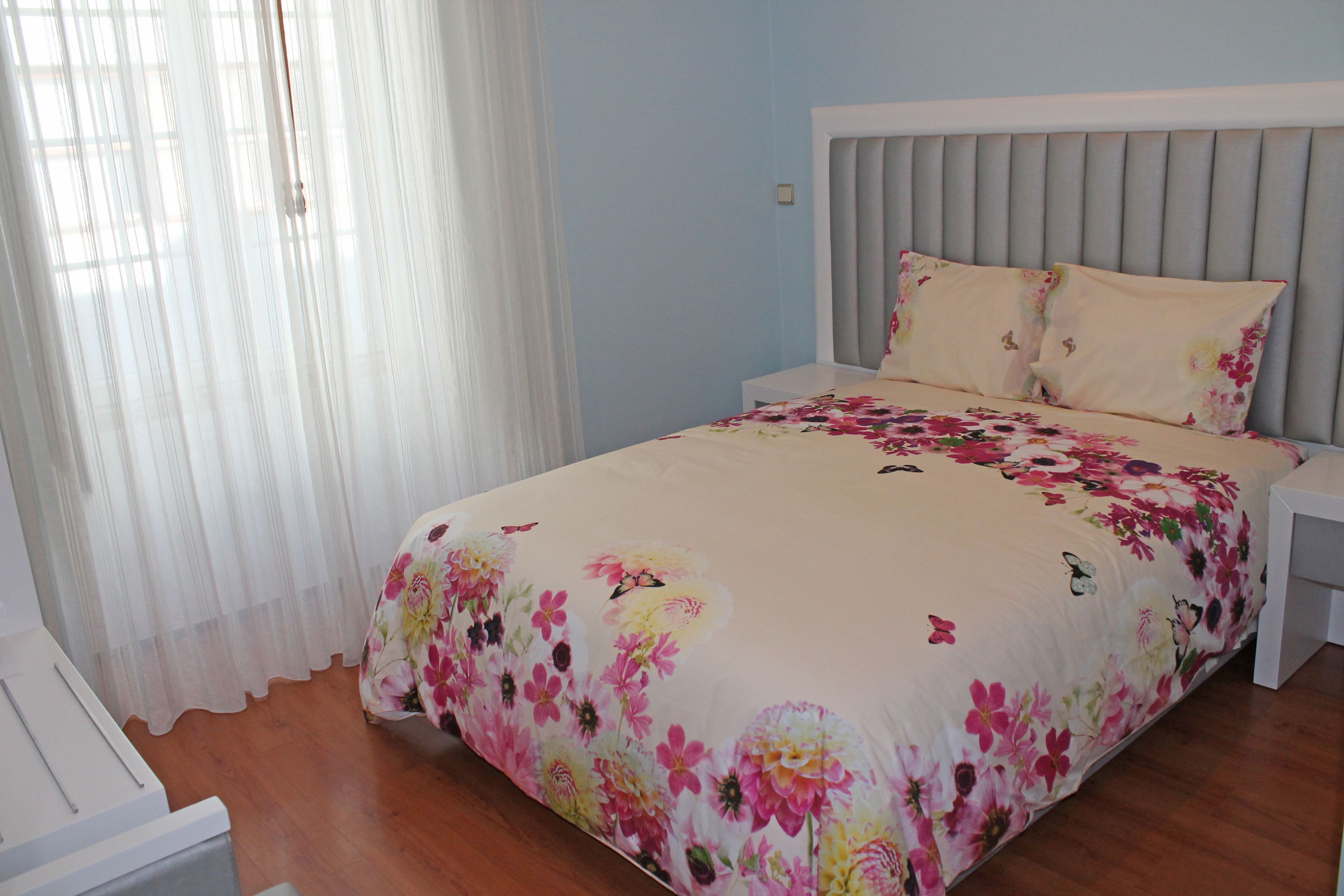 A bedroom with a bed, nightstands, a window with sheer curtains, and a wooden floor.