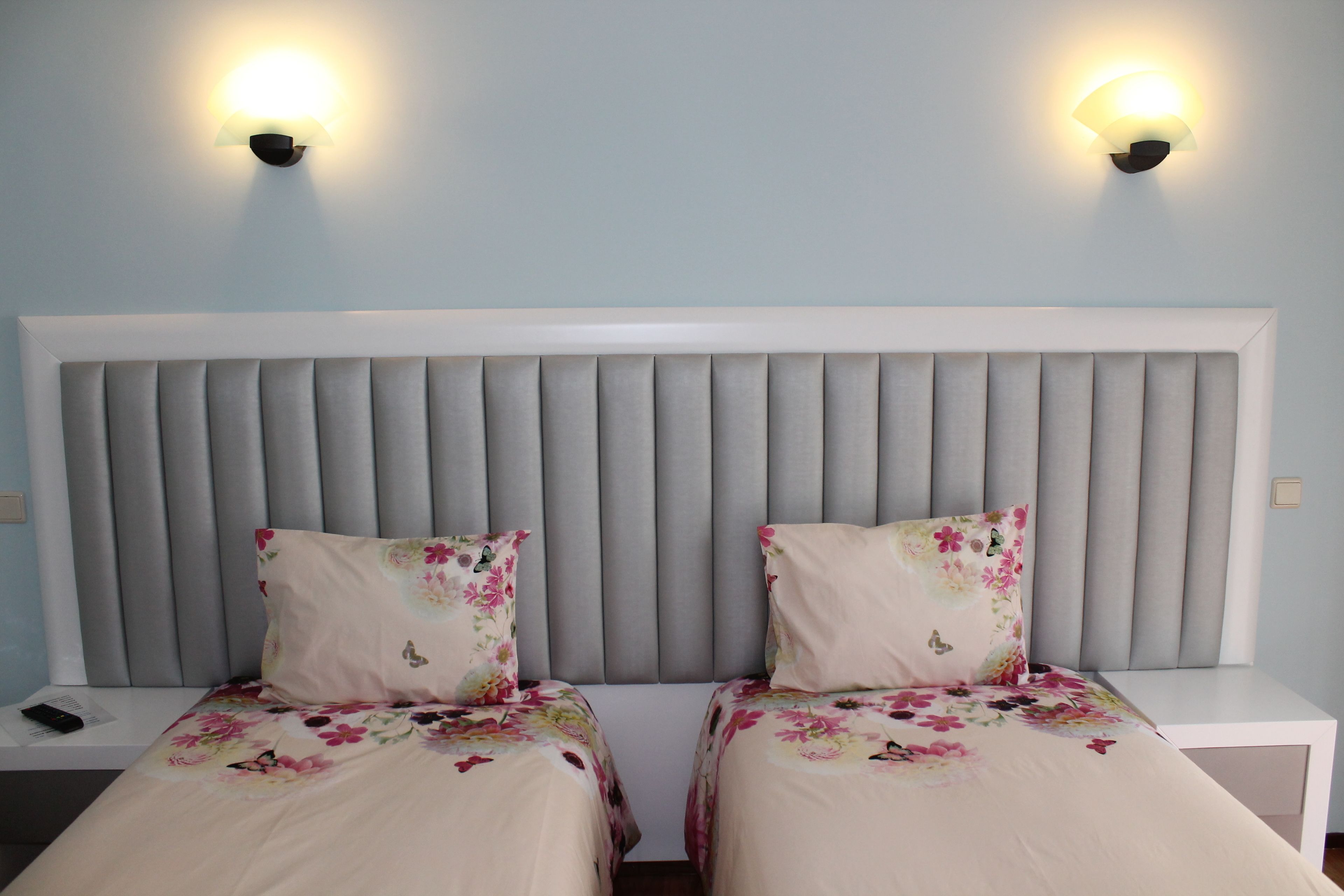 Uma cama de casal com cabeceira estofada cinza, duas mesas de cabeceira e duas luminárias fixadas na parede.