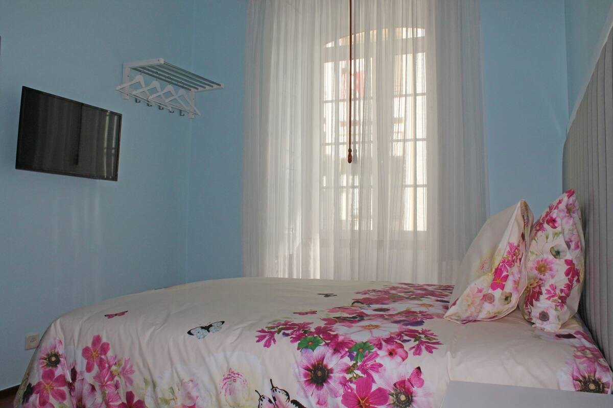 Een slaapkamer met een bed, een bloemenprint beddegoed, witte gordijnen en een vastgezette airconditioning.