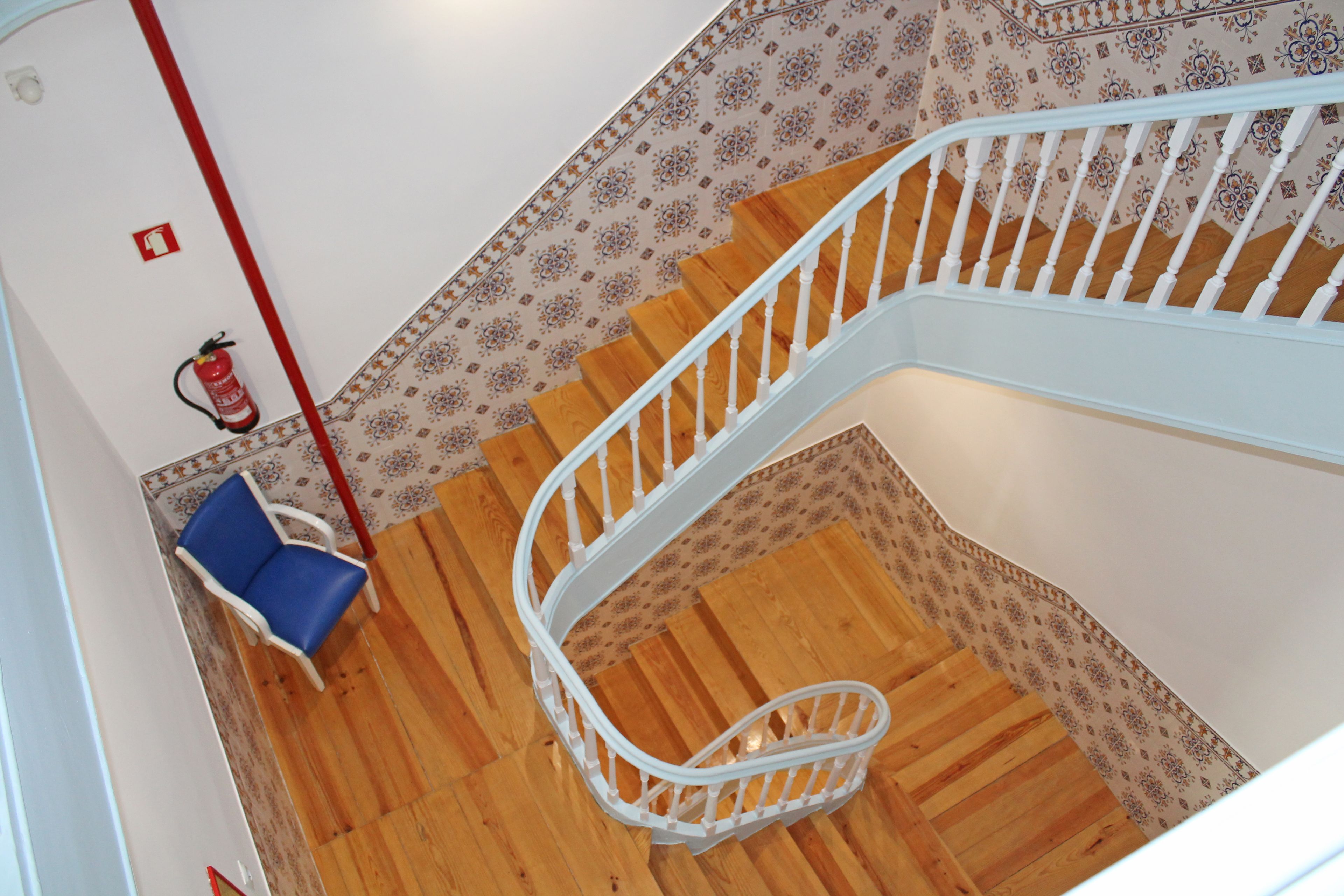Een wenteltrap met houten treden en witte balustrades, leidend naar een plateau met een blauwe stoel en een brandblusser aan de muur bevestigd.