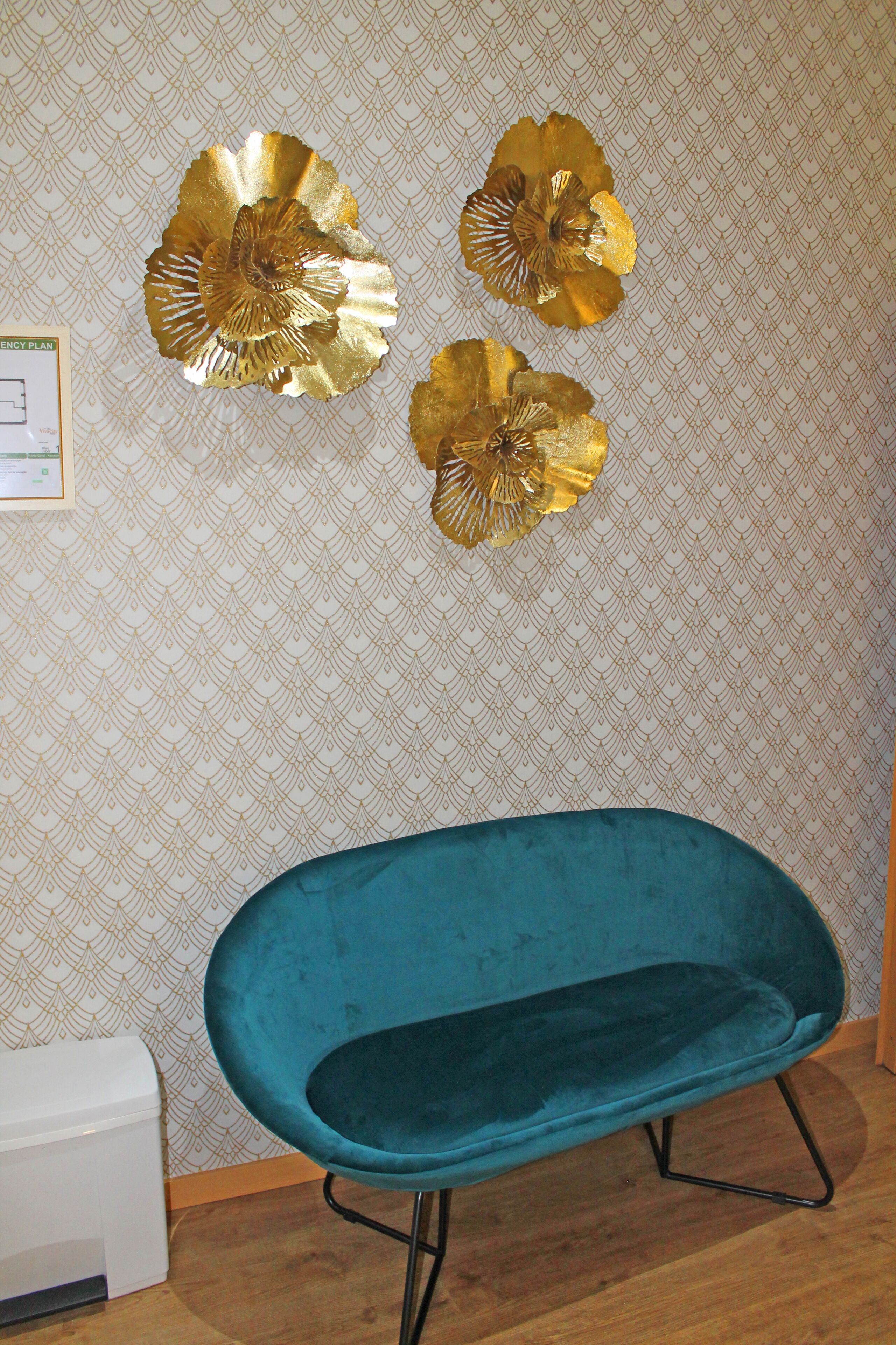 A teal chair with a metal frame, a patterned wallpaper, and three gold decorative pieces on the wall.