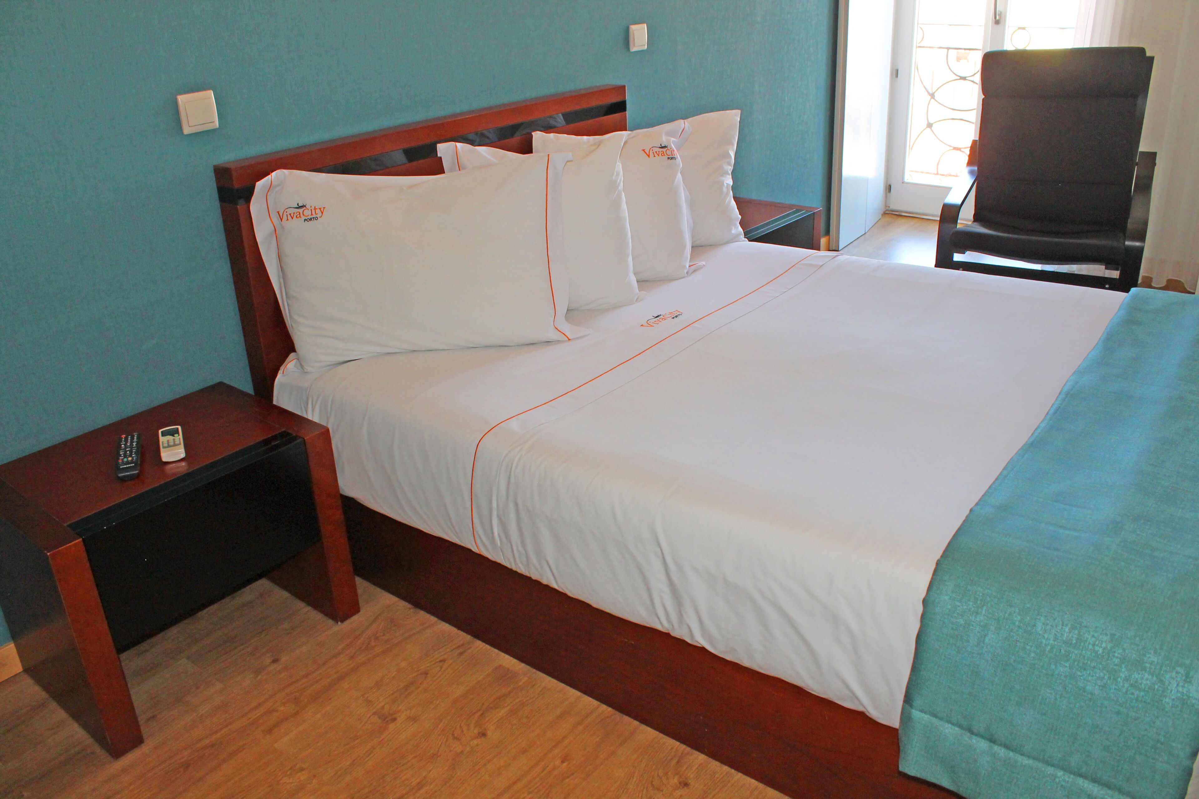 Una cama bien hecha con sábanas blancas y ribetes naranja, una mesita de noche de madera con un control remoto, y una silla en una habitación con paredes color verde azulado.