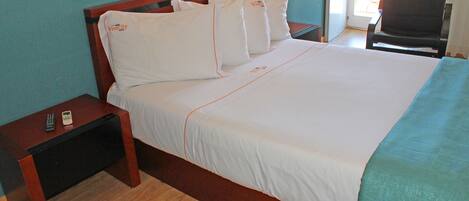 Una cama bien hecha con sábanas blancas y ribetes naranja, una mesita de noche de madera con un control remoto, y una silla en una habitación con paredes color verde azulado.