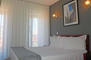 A hotel room with a large bed, white bedding, a framed picture on the wall, and a window with sheer curtains.