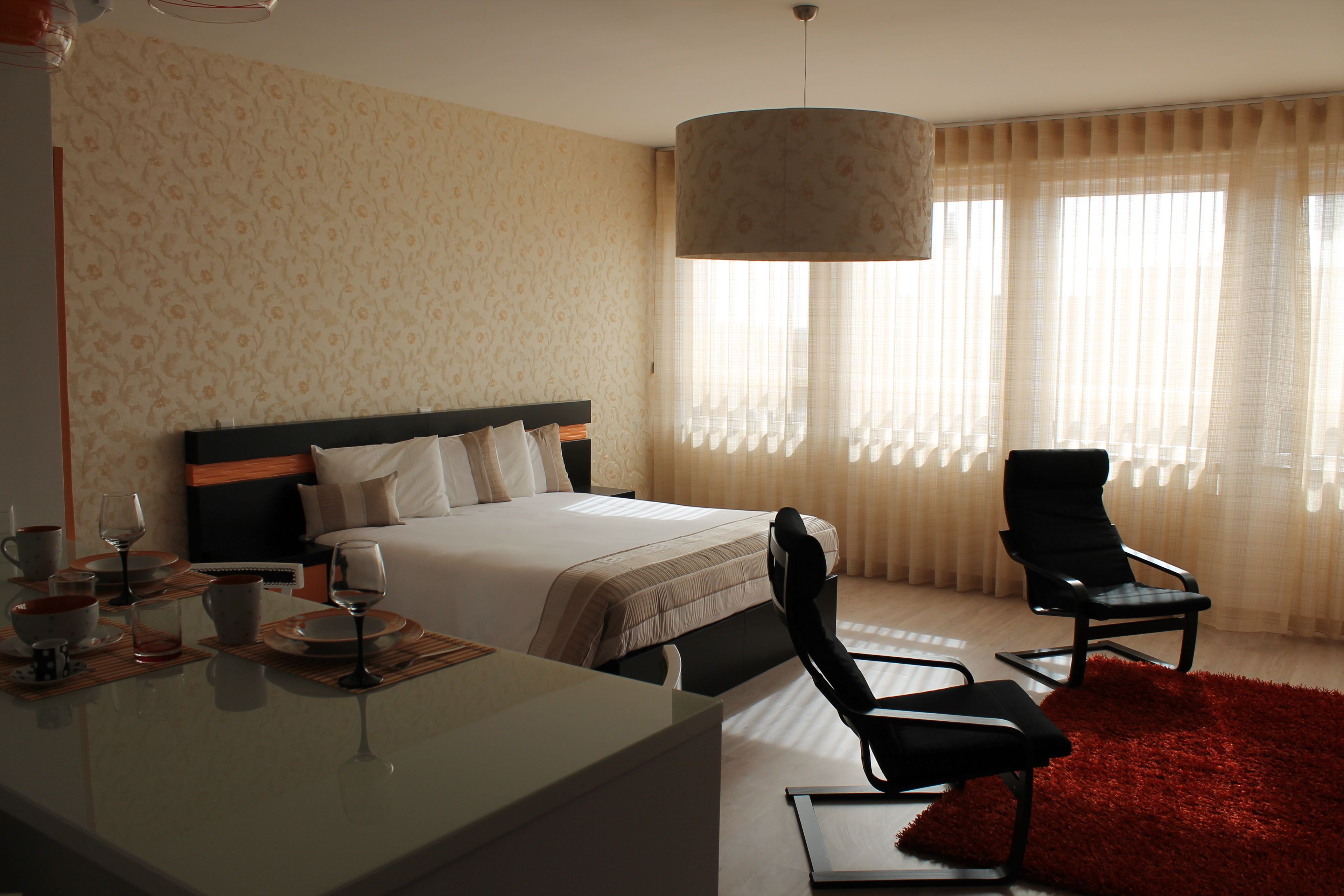 A hotel room with a bed, a desk with a chair, a red rug, and a large window with sheer curtains.