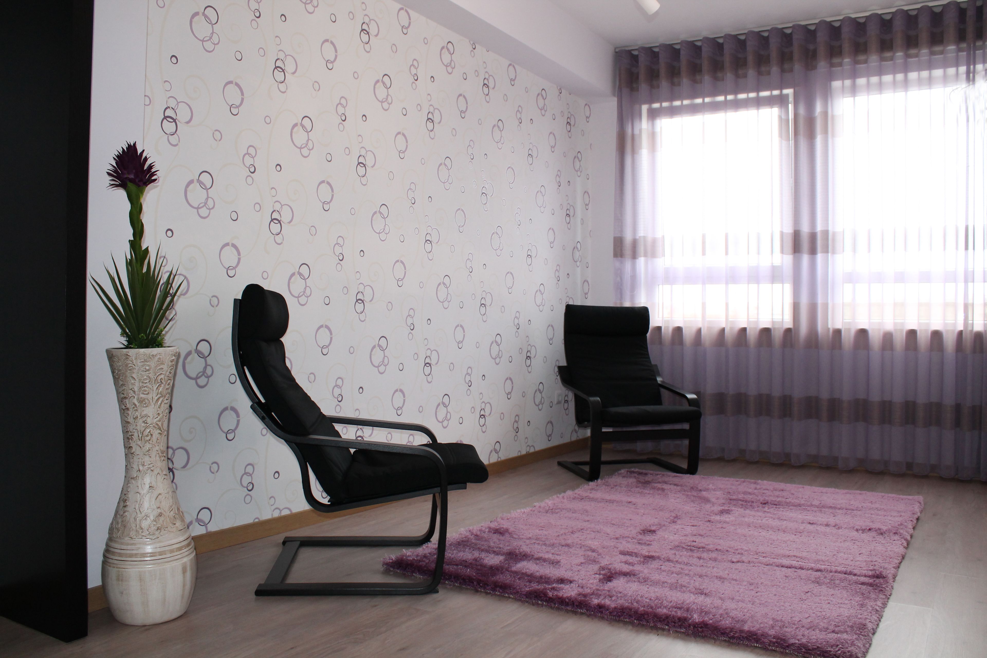A modern living room with a patterned wallpaper, a black chair, a small table, and a large vase with a plant.