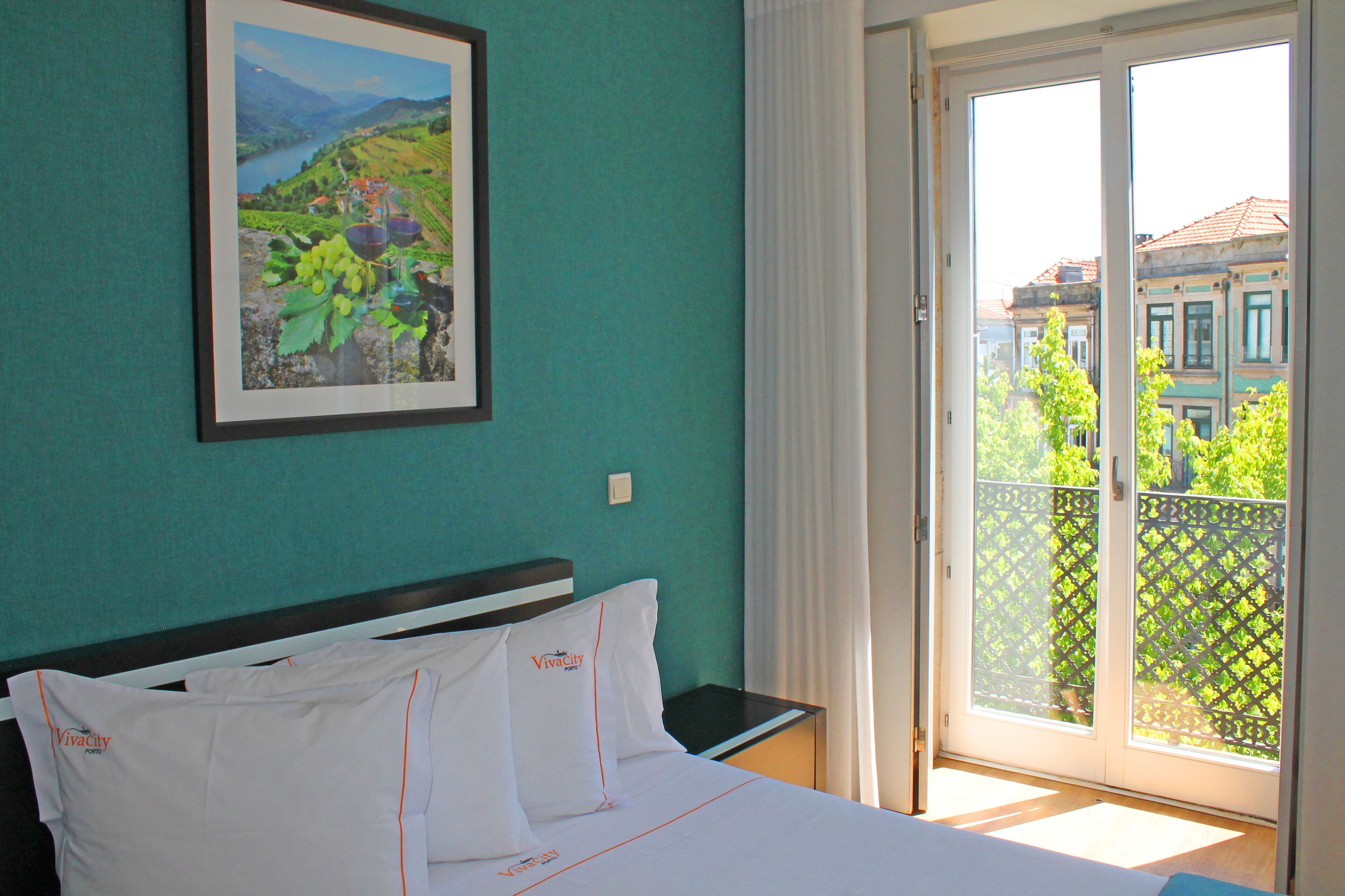 Une chambre d’hôtel avec un lit, un tableau encadré représentant un vignoble et un balcon offrant une vue sur des bâtiments et de la verdure.