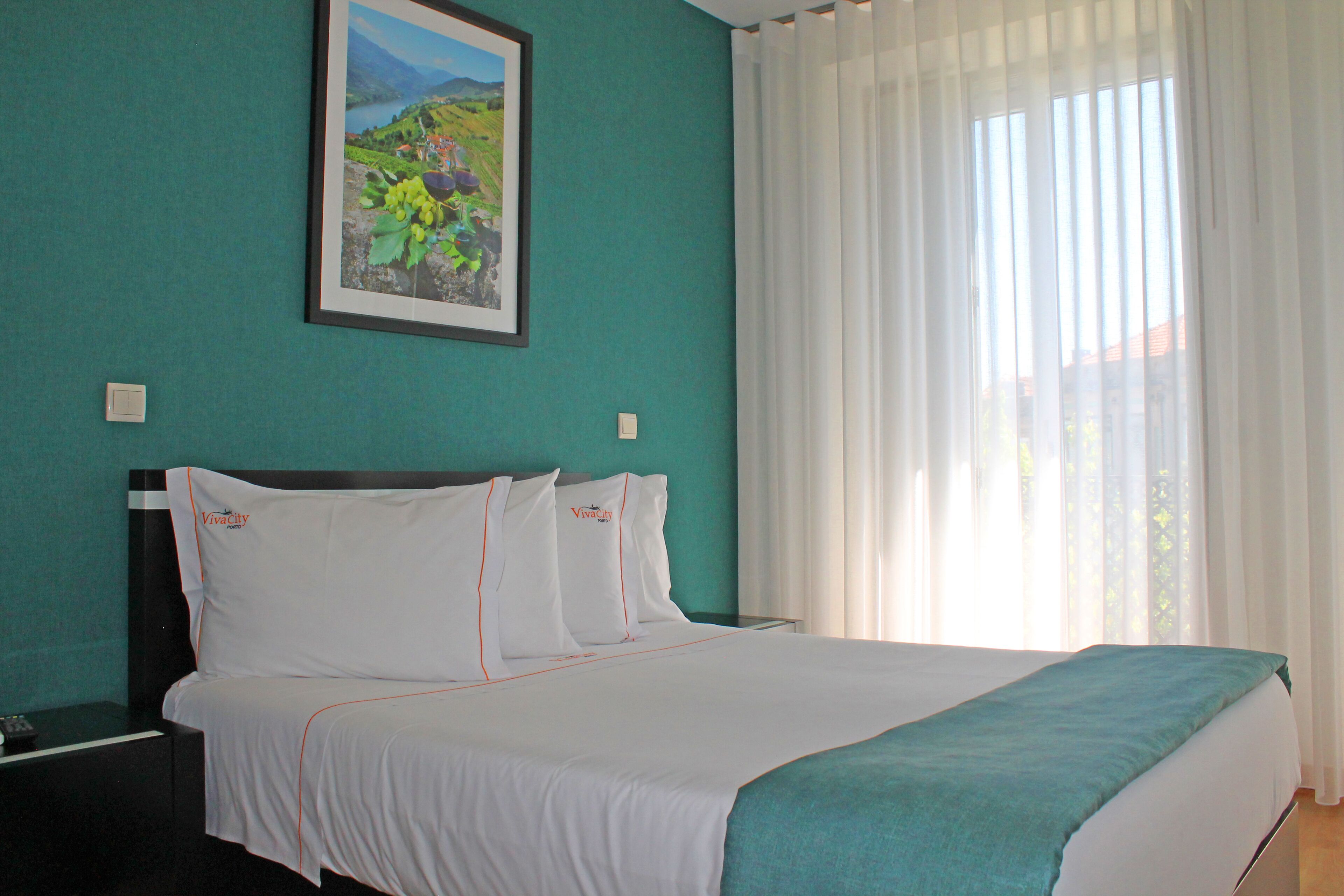 A neatly made bed with white linens and a teal throw blanket, a framed landscape painting on the teal wall, and a window with sheer white curtains.