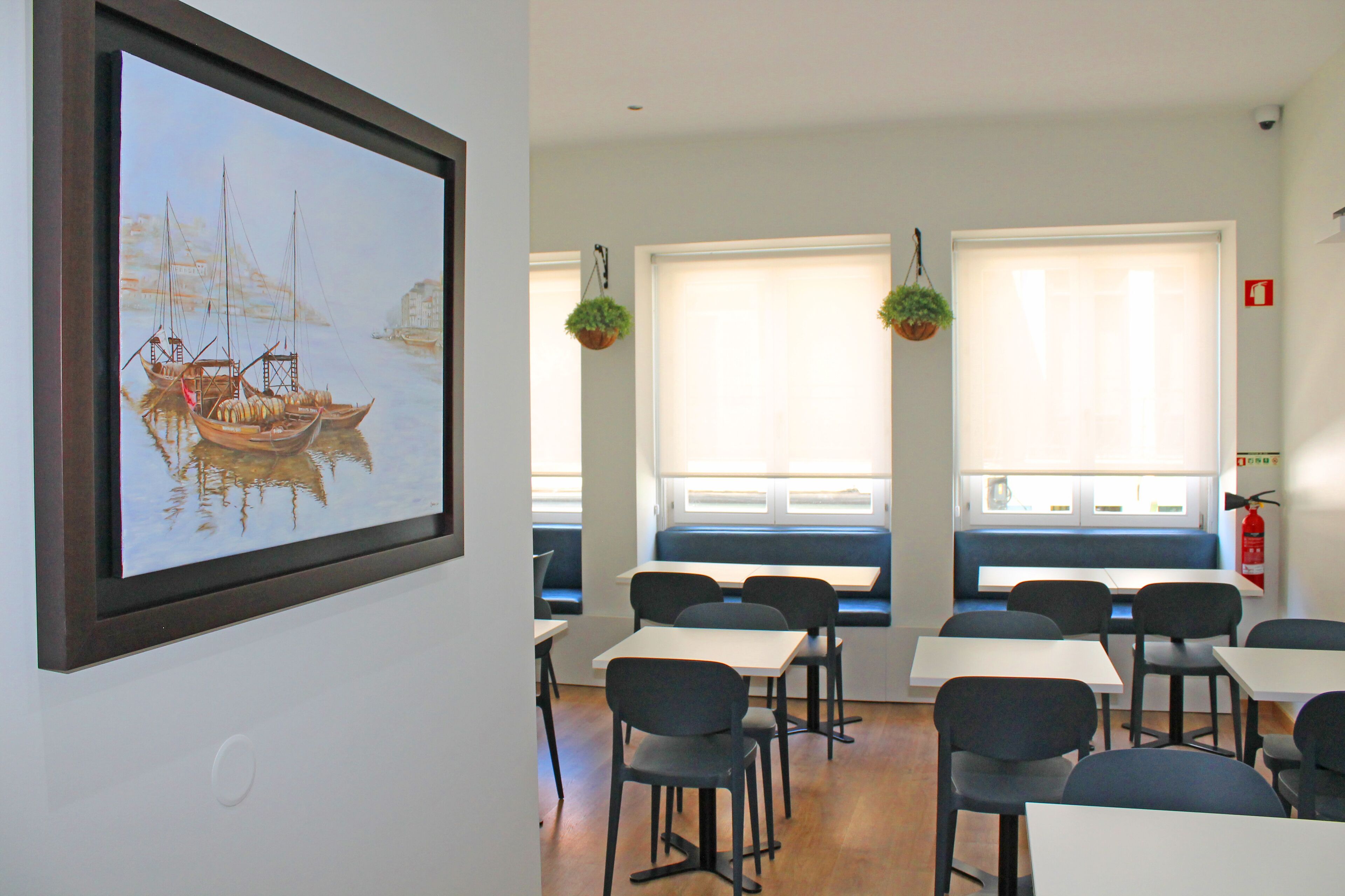 A dining area with tables and chairs, a painting of boats on the wall, and windows with hanging plants.