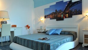 Um quarto com cama, escrivaninha, cadeira, mesa de cabeceira, luminária e uma obra de arte na parede.