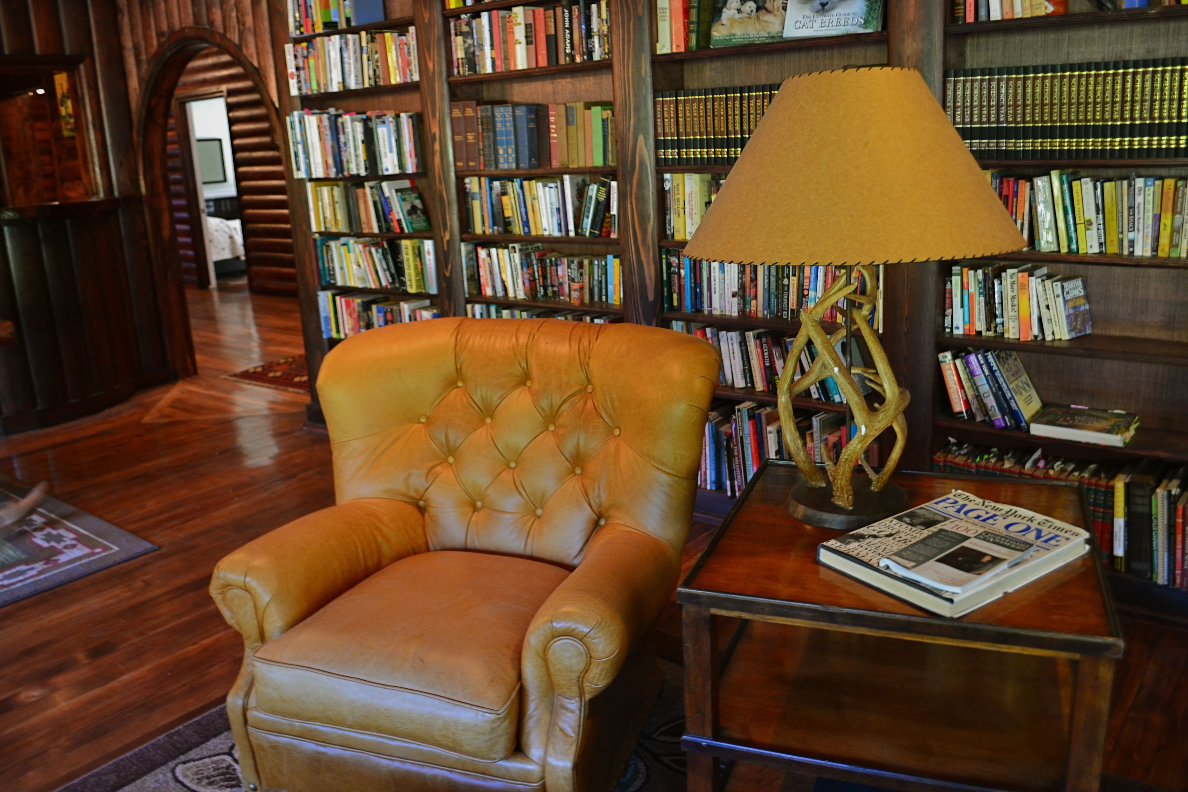Un fauteuil en cuir dans une bibliothèque, avec des étagères remplies de livres, une lampe et un journal sur une petite table d’appoint.