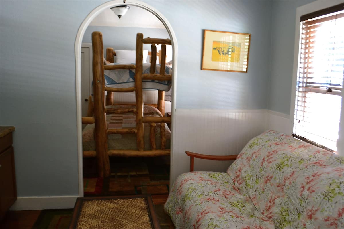 Une chambre avec un lit-échelle en bois, une couvre-lit à motifs floraux, un tableau encadré au mur et une fenêtre avec des stores.