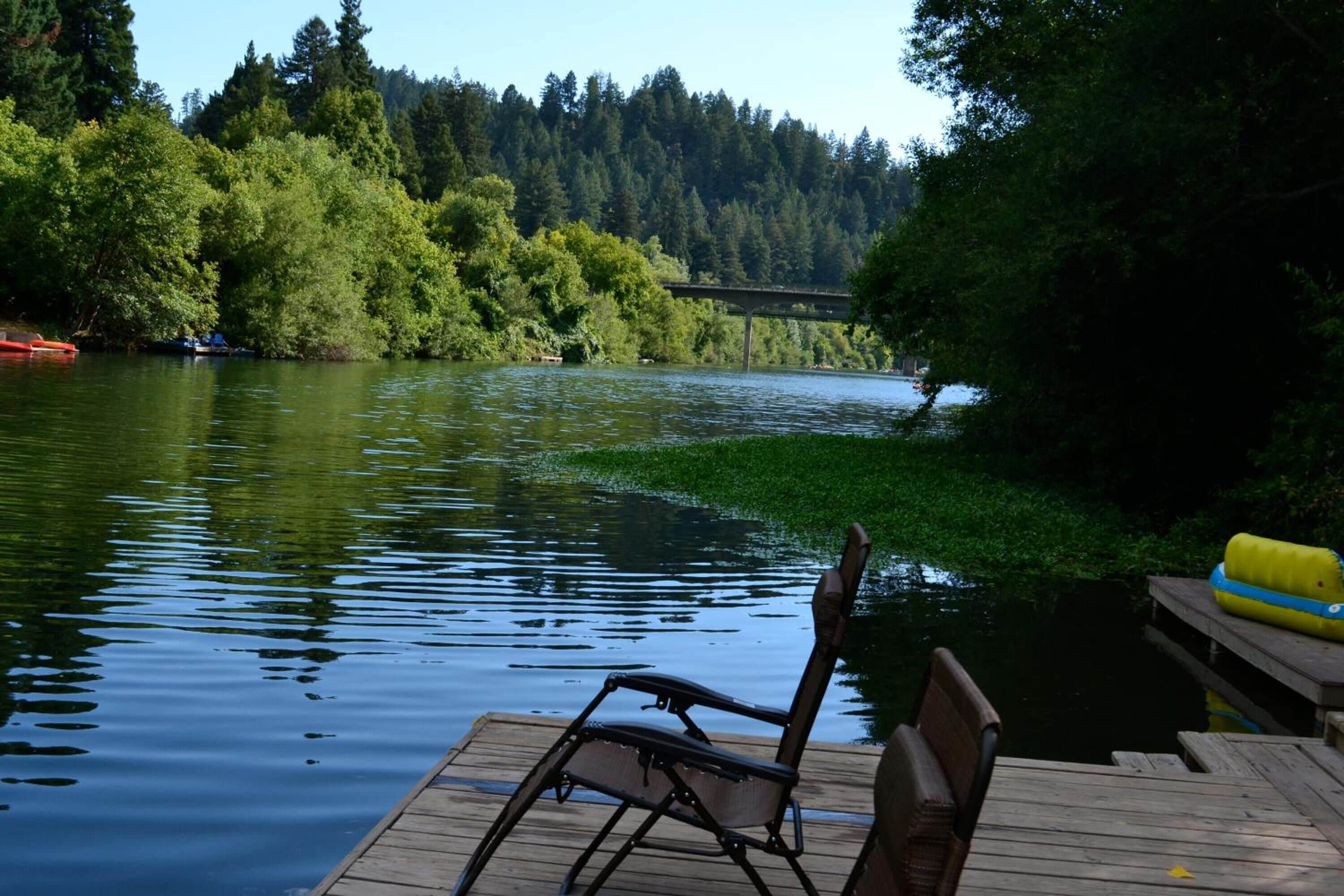 A wooden dock with a chair and a table, overlooking a calm lake with a forested area and a bridge in the background.
