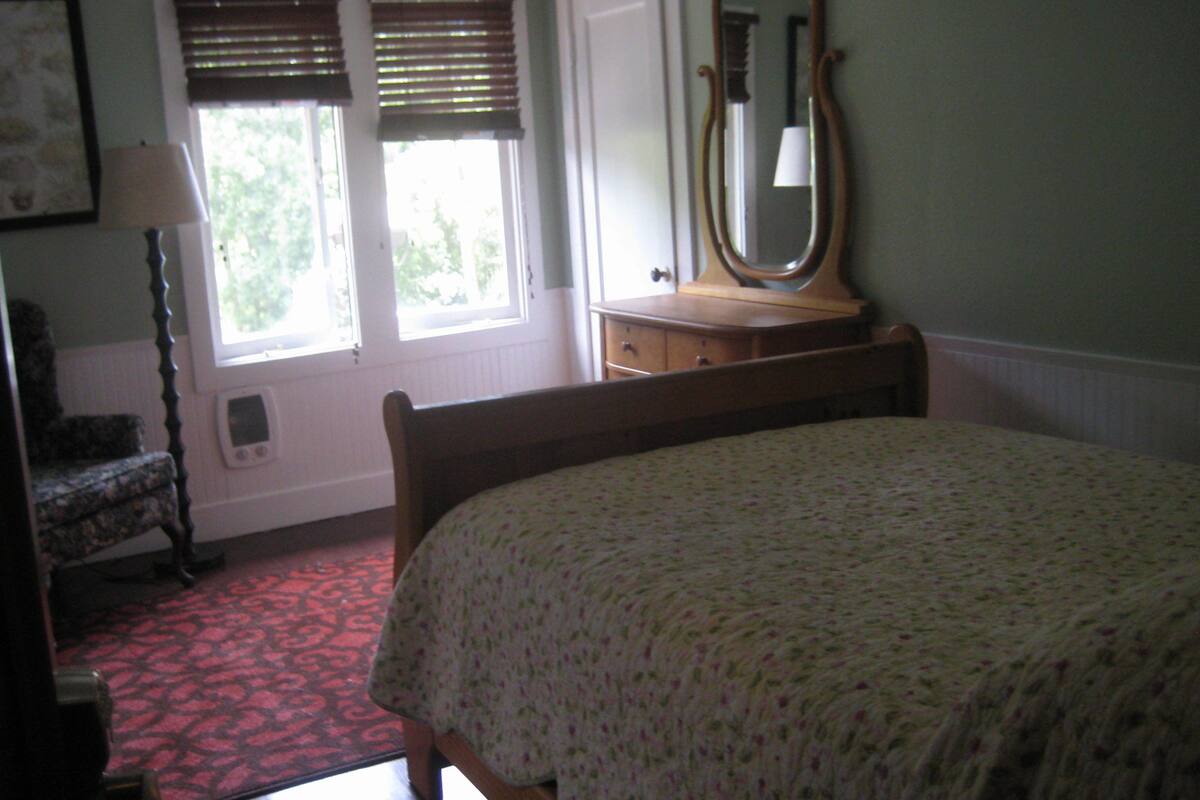 Une chambre à coucher comprenant un lit, une commode, une chaise, un miroir et une fenêtre avec des stores.