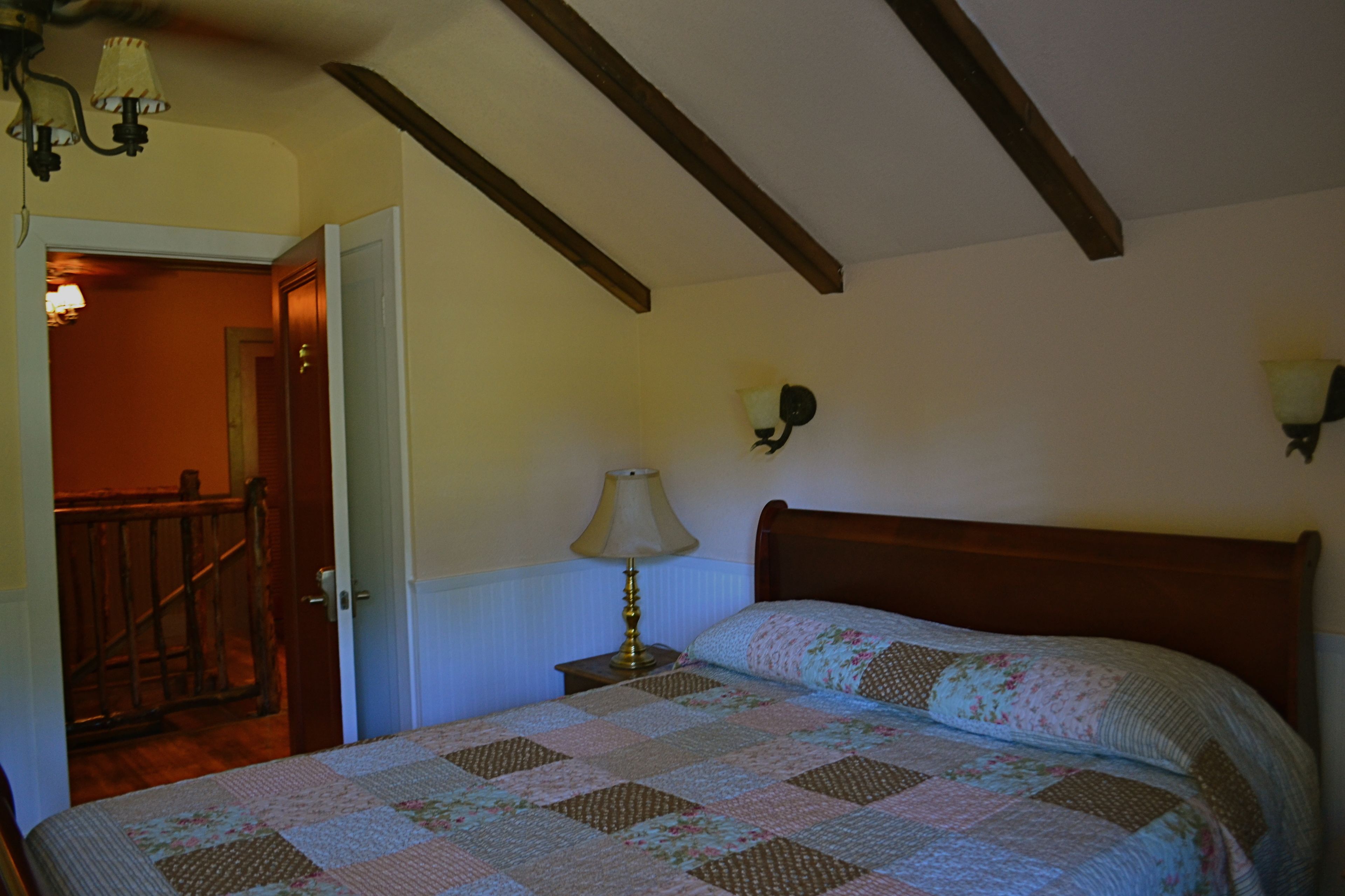 Une chambre avec un plafond en bois, un lit recouvert d’une courtepointe à motifs floraux, une table de chevet avec une lampe et un luminaire fixé au mur.