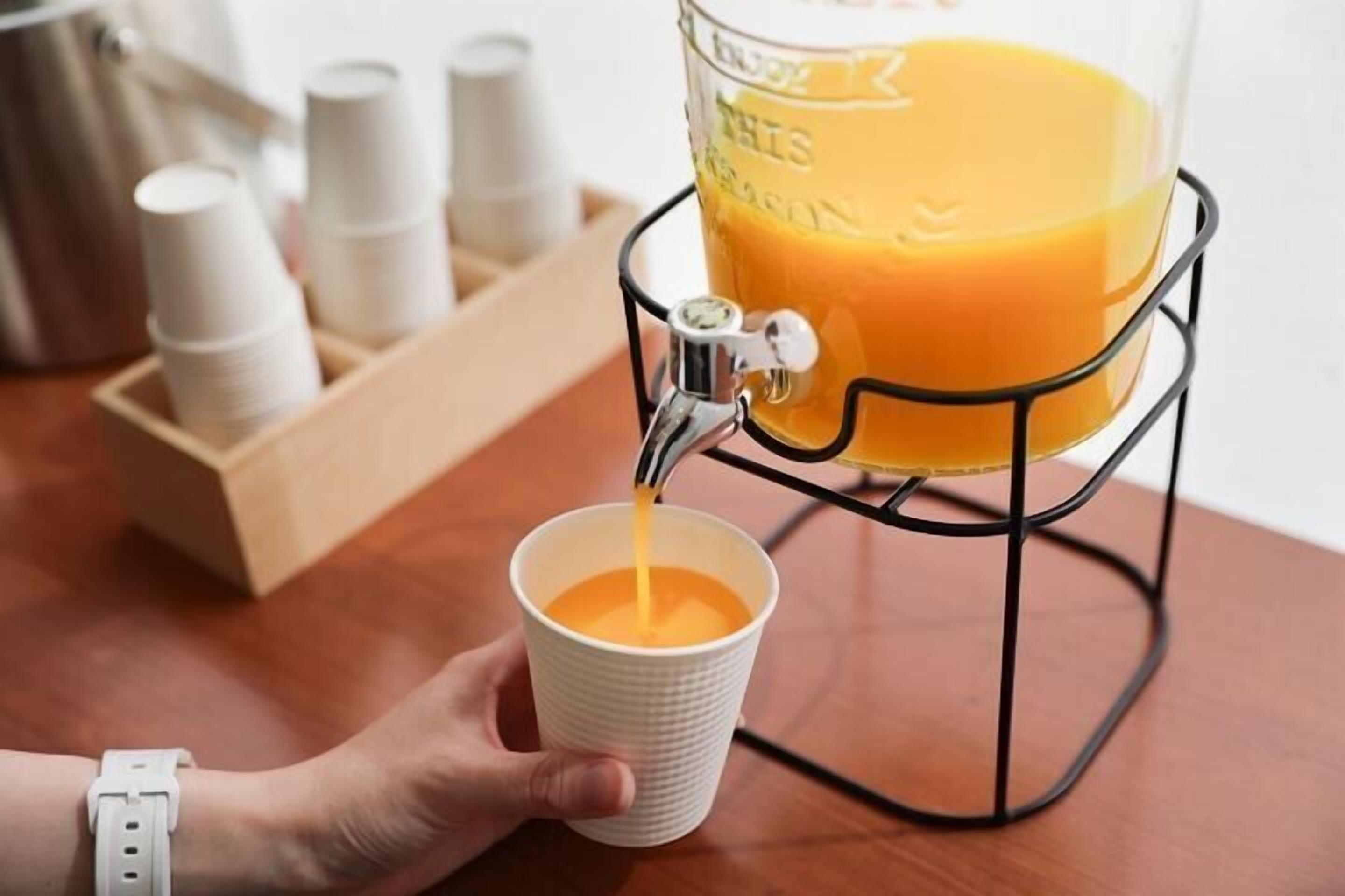 A hand holding a cup under a juice dispenser, with a container of orange juice and paper cups in the background.