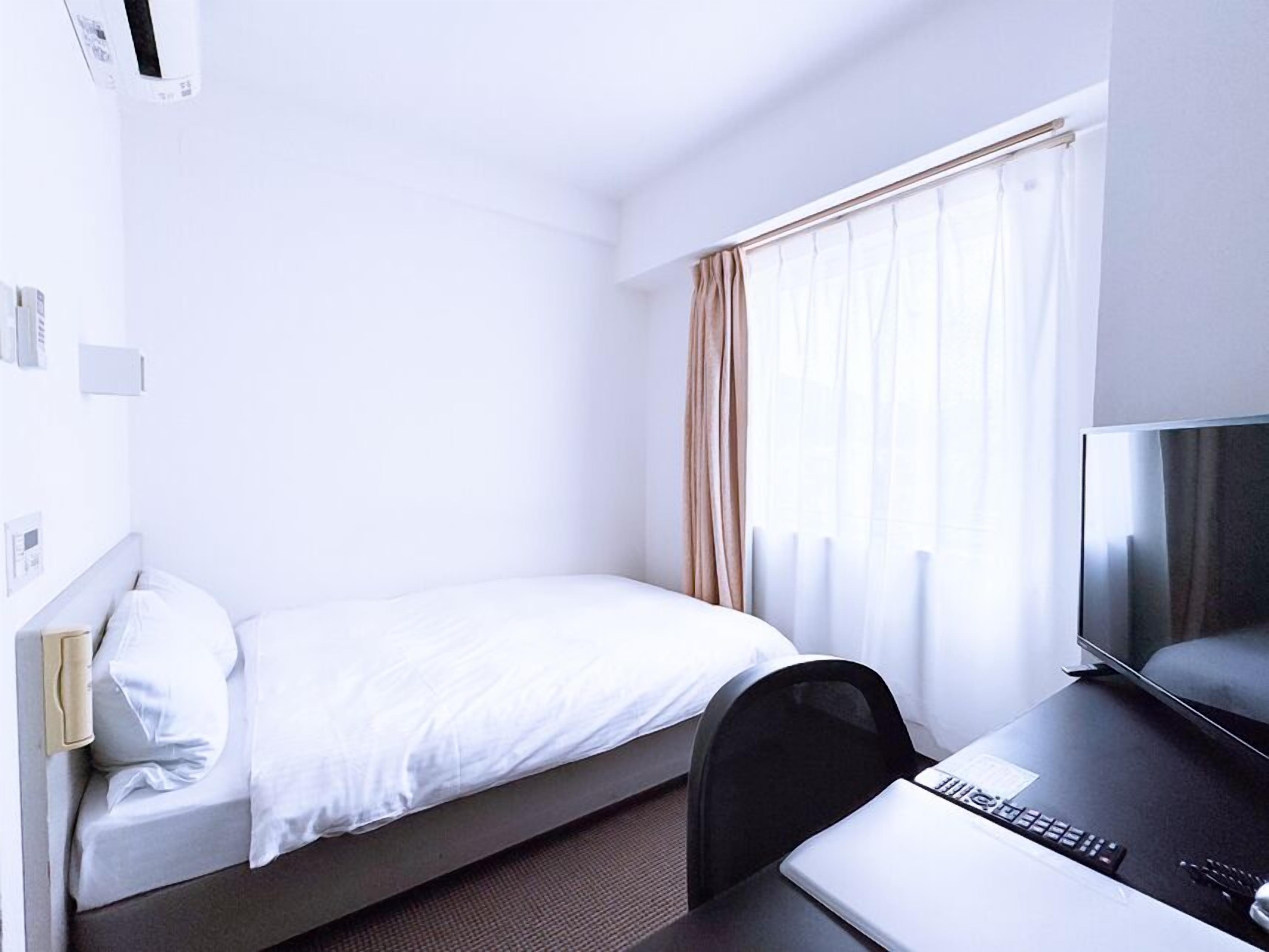 Una cama individual con colchón y almohadas blancos, un escritorio con una computadora portátil y un control remoto, un televisor y una ventana con cortinas.