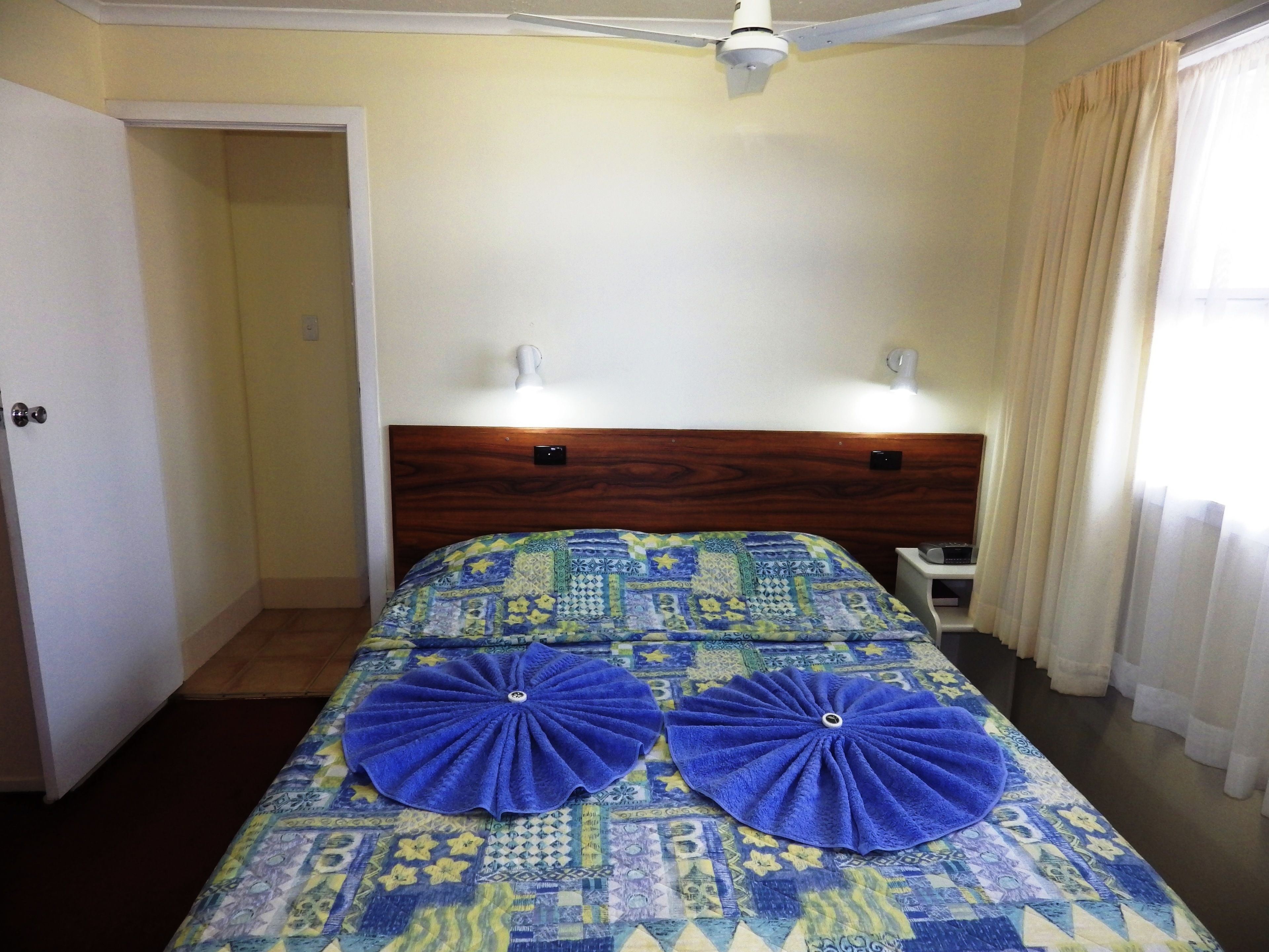 A hotel room with a wooden headboard, two wall-mounted lights, a patterned bedspread, and a door leading to another room.