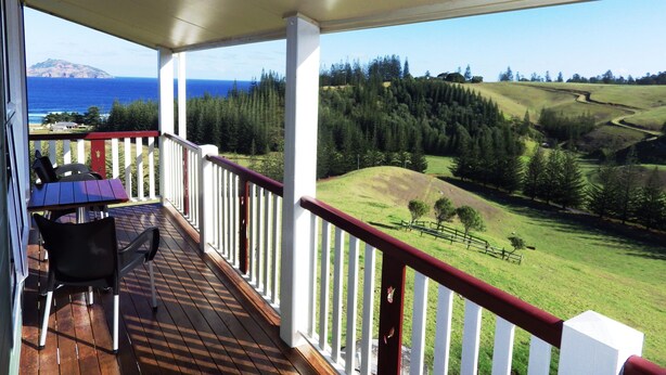 A covered balcony with a table and chairs, overlooking a scenic landscape with a view of the ocean and hills. - Panorama Seaside Apartments (Norfolk Island)