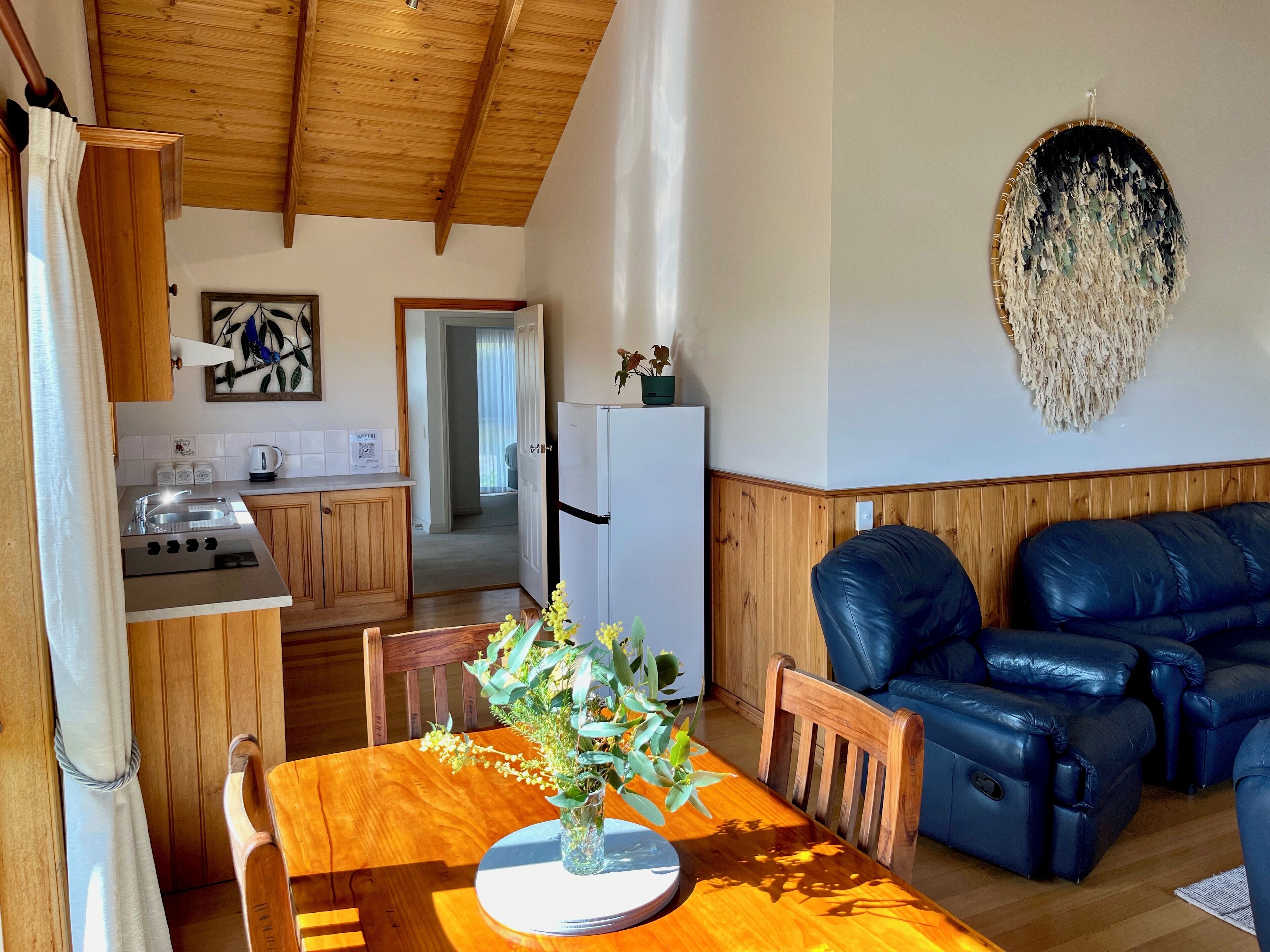 A modern kitchen with wooden cabinets, a dining table with a vase of flowers, and a black leather sofa in a living area.