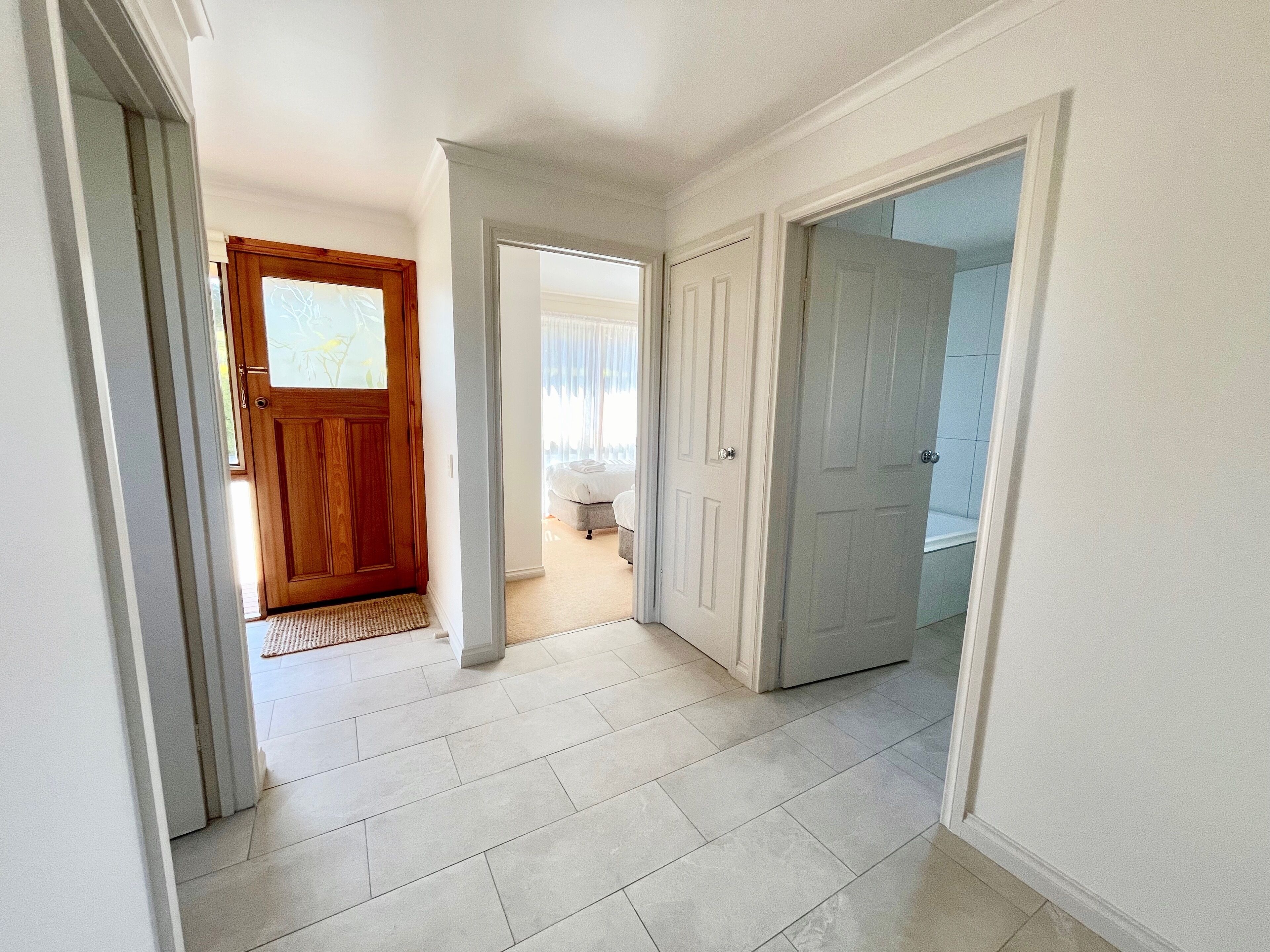 A hallway with a wooden door, a white door, and a view into a bedroom with a bed and curtains.