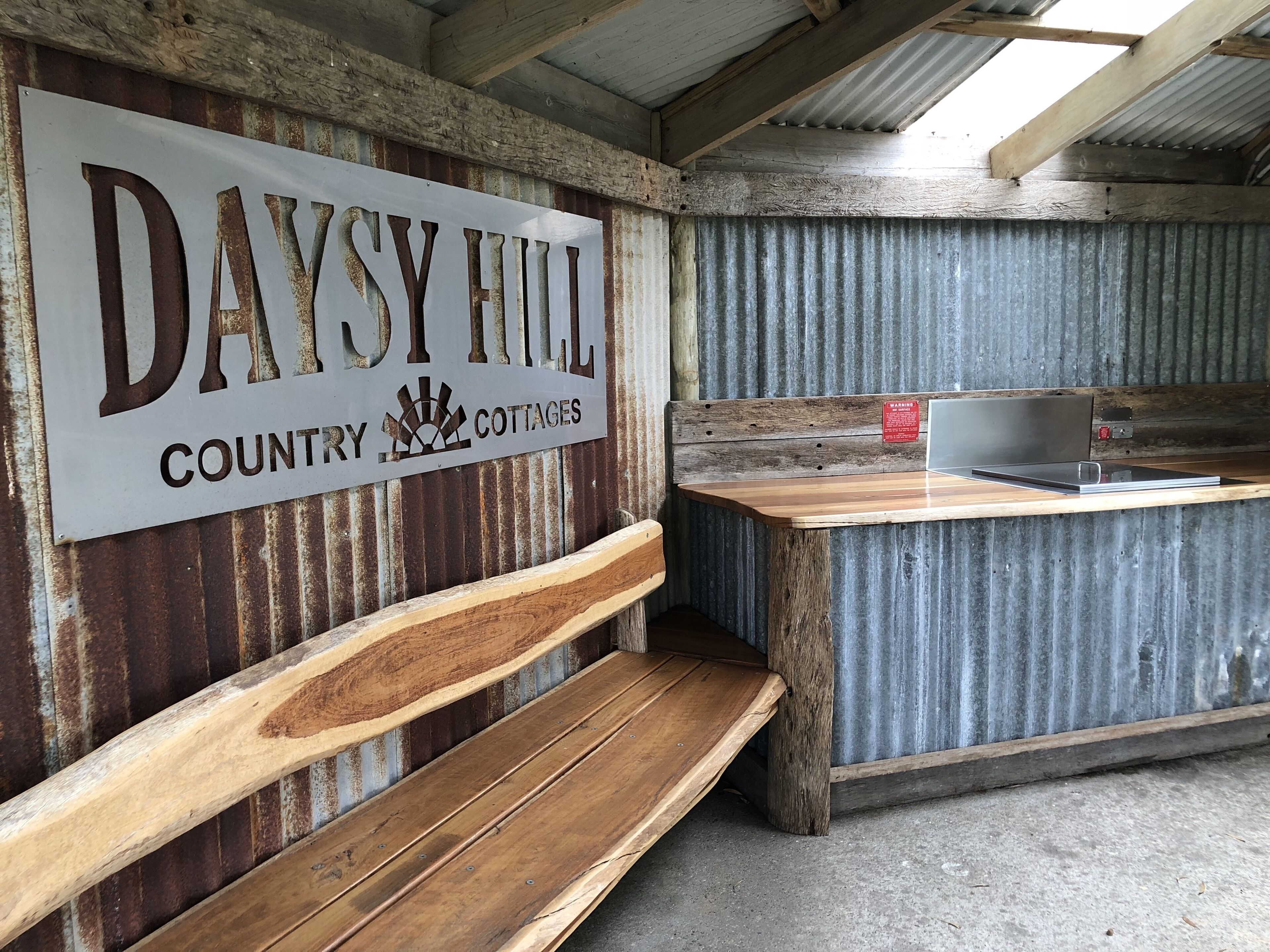 Un interno rustico con una panca di legno, un cartello metallico con la scritta “DAYSY HILL COUNTRY COTTAGES” e un bancone in legno con un portatile.