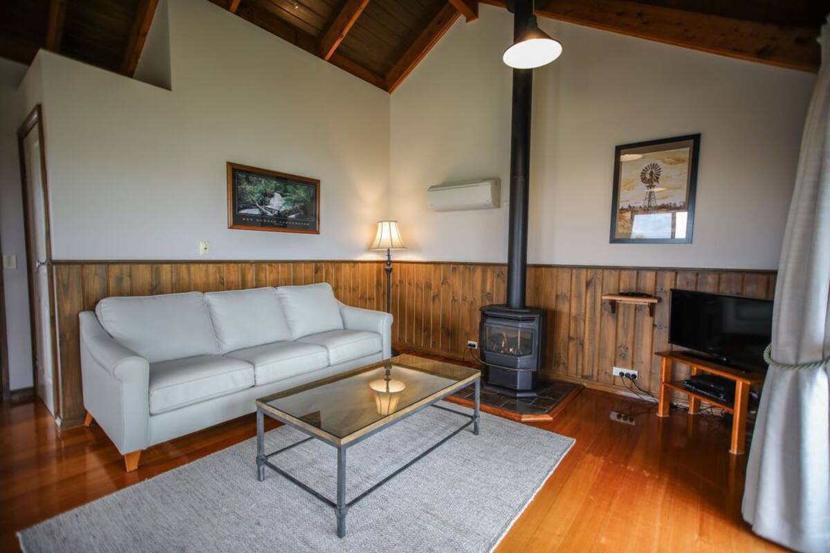 Una sala de estar acogedora con un sofá blanco, una mesa de centro de vidrio, una estufa de leña, un televisor sobre un soporte de madera y cuadros enmarcados en las paredes.