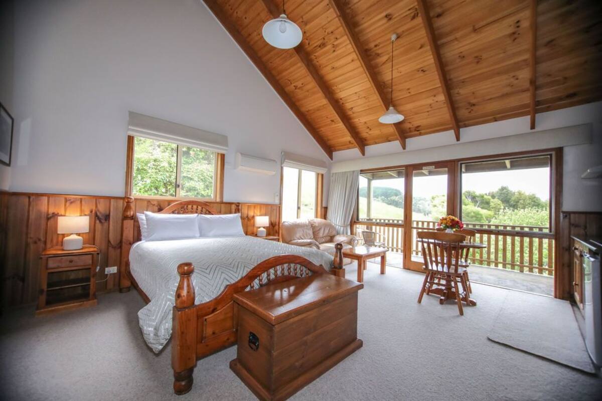 Un dormitorio amplio con techo de madera, una cama grande, un cofre de madera, un escritorio con una silla, y un balcón con vistas a zonas verdes.