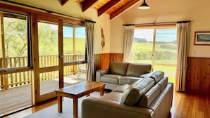 Uma sala de estar aconchegante com um sofá, uma mesa pequena e vista para um deck de madeira e vegetação lá fora.