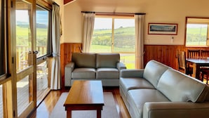 Uma sala de estar espaçosa com um sofá grande, uma mesa de centro de madeira e um espaço de jantar com mesa e cadeiras de madeira. Grandes janelas proporcionam vista para o campo.