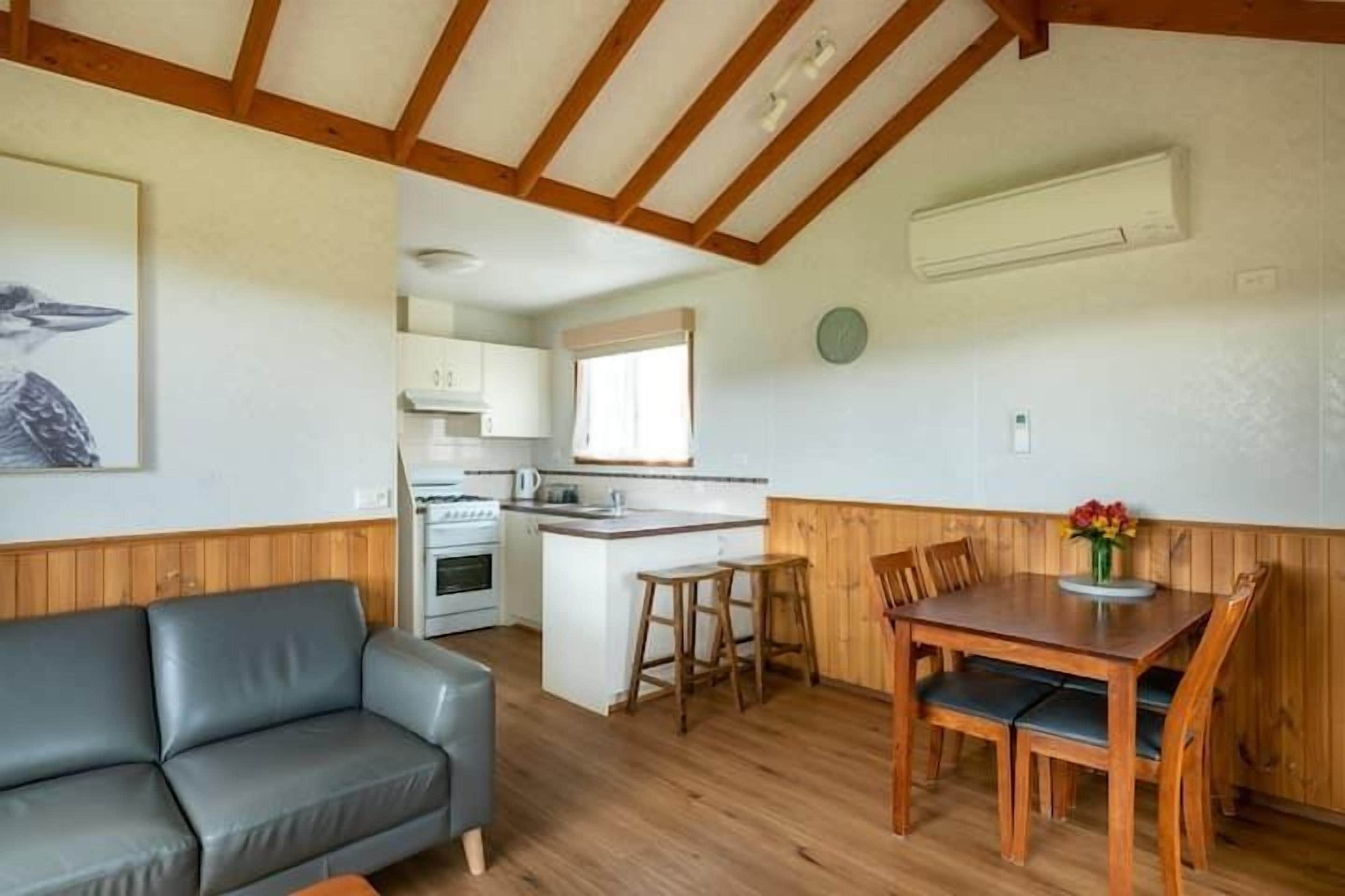 Ett mysigt rum med ett träpanelat tak, en grå soffa, ett träbord med stolar och ett köksutrymme i bakgrunden.