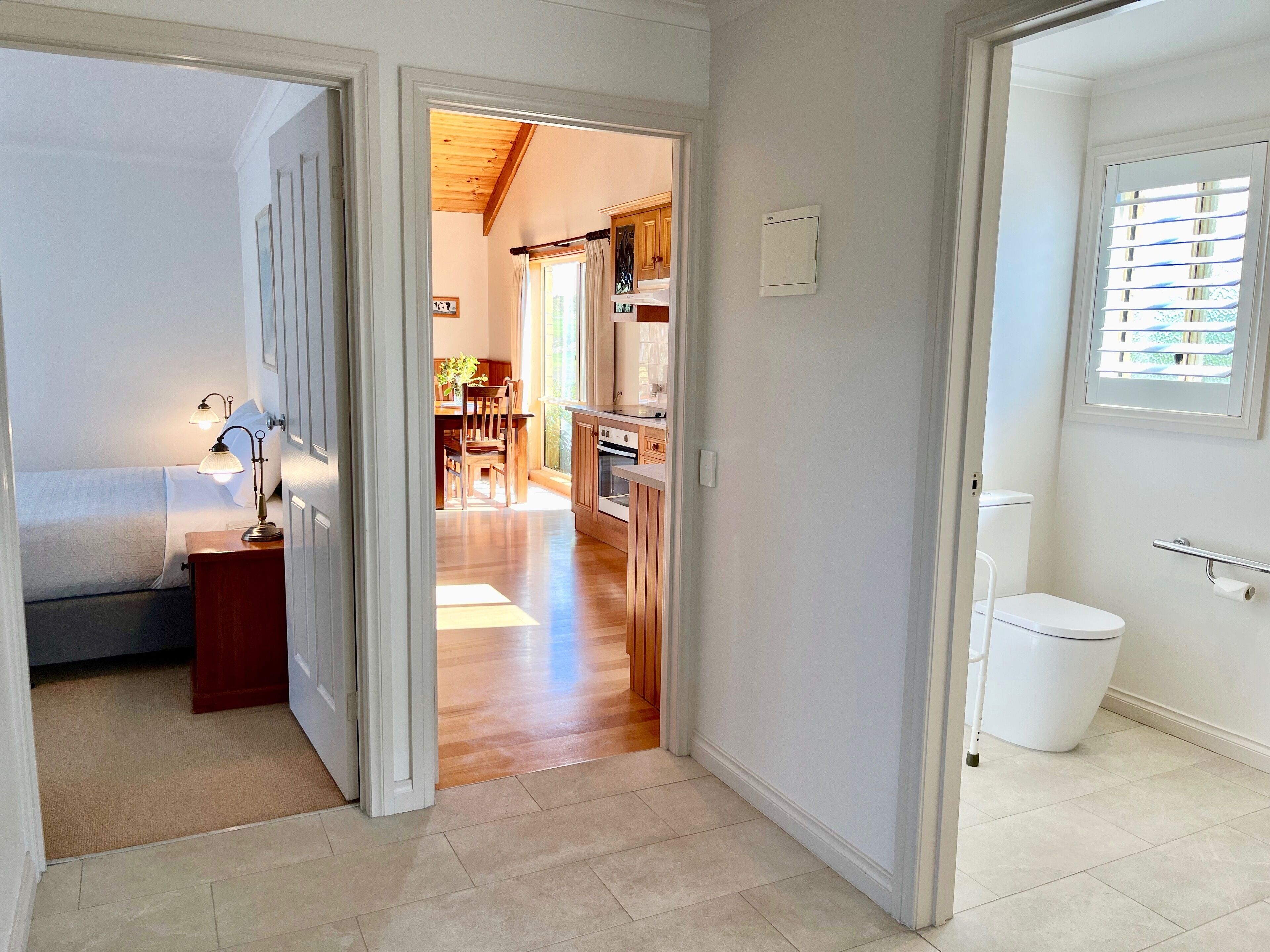 Un dormitorio con cama, mesita de noche y lámpara, que da paso a una cocina con armarios de madera y una mesa de comedor.
