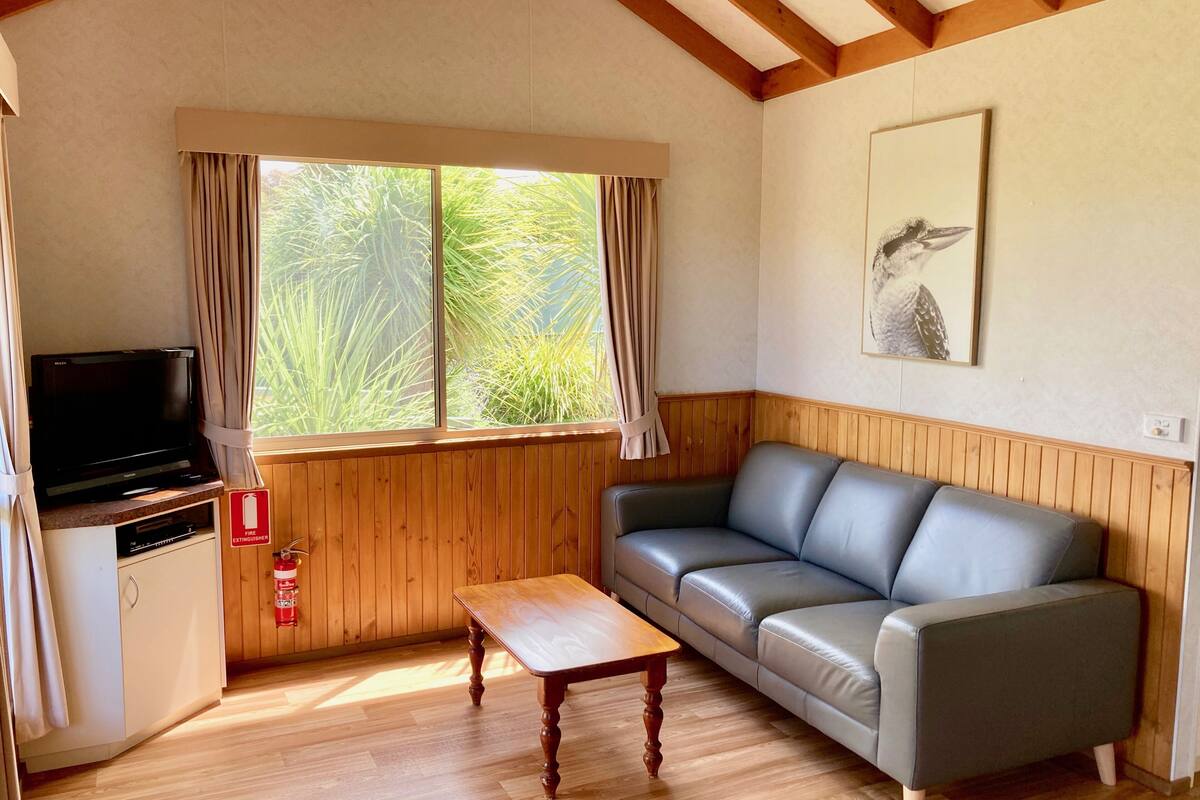 Una habitación con piso de madera, un sofá de cuero gris, una mesa de centro de madera, un televisor sobre un mueble bajo y una ventana con cortinas.
