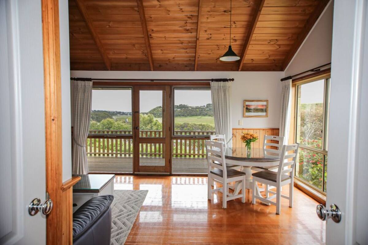 Una habitación con techo de madera, un ventanal corredero que da a una terraza exterior, una mesa de comedor con sillas y un banco acolchado.