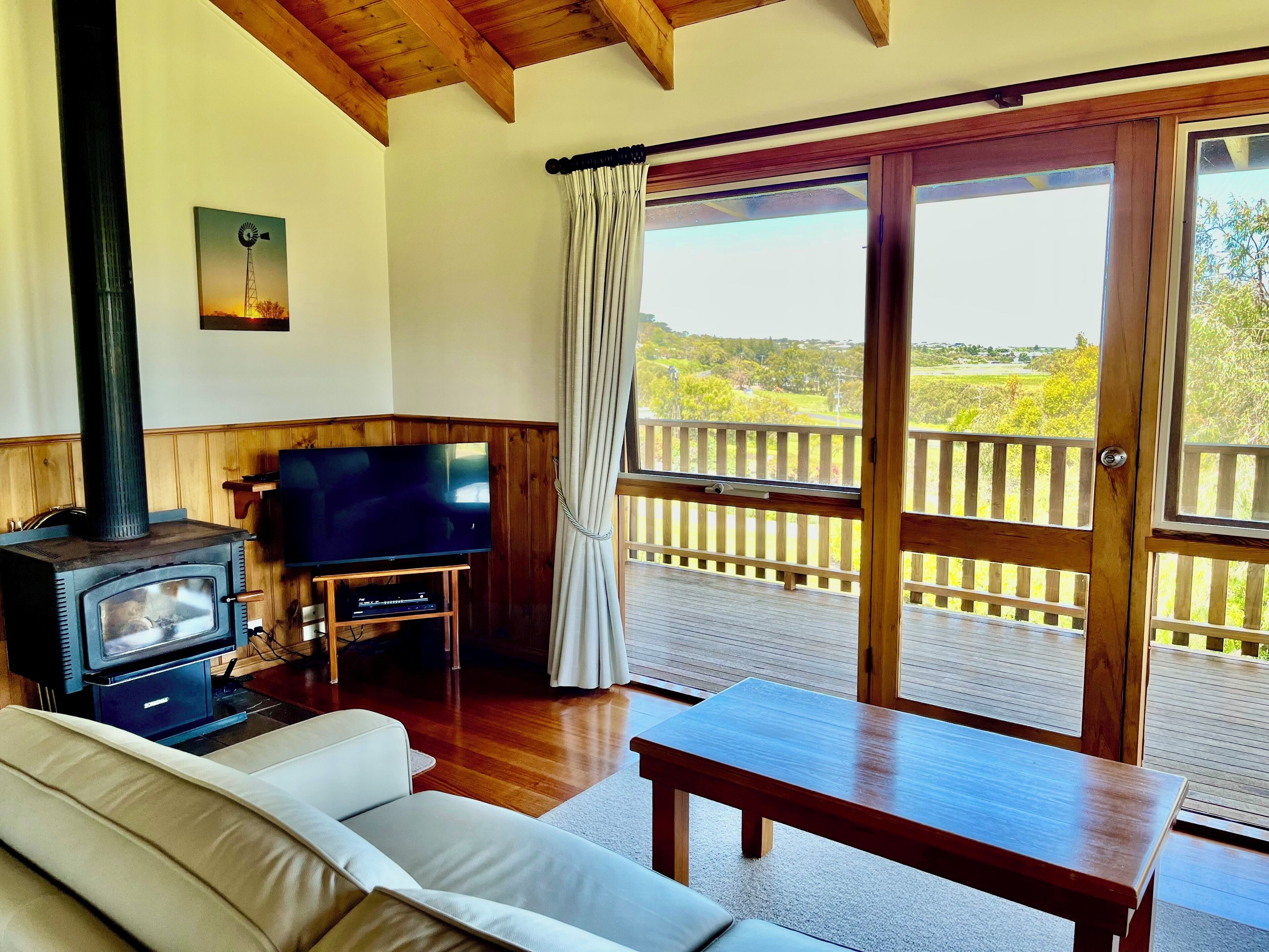 A cozy living room with a television, a sofa, a wooden table, and a large window with a view of greenery.