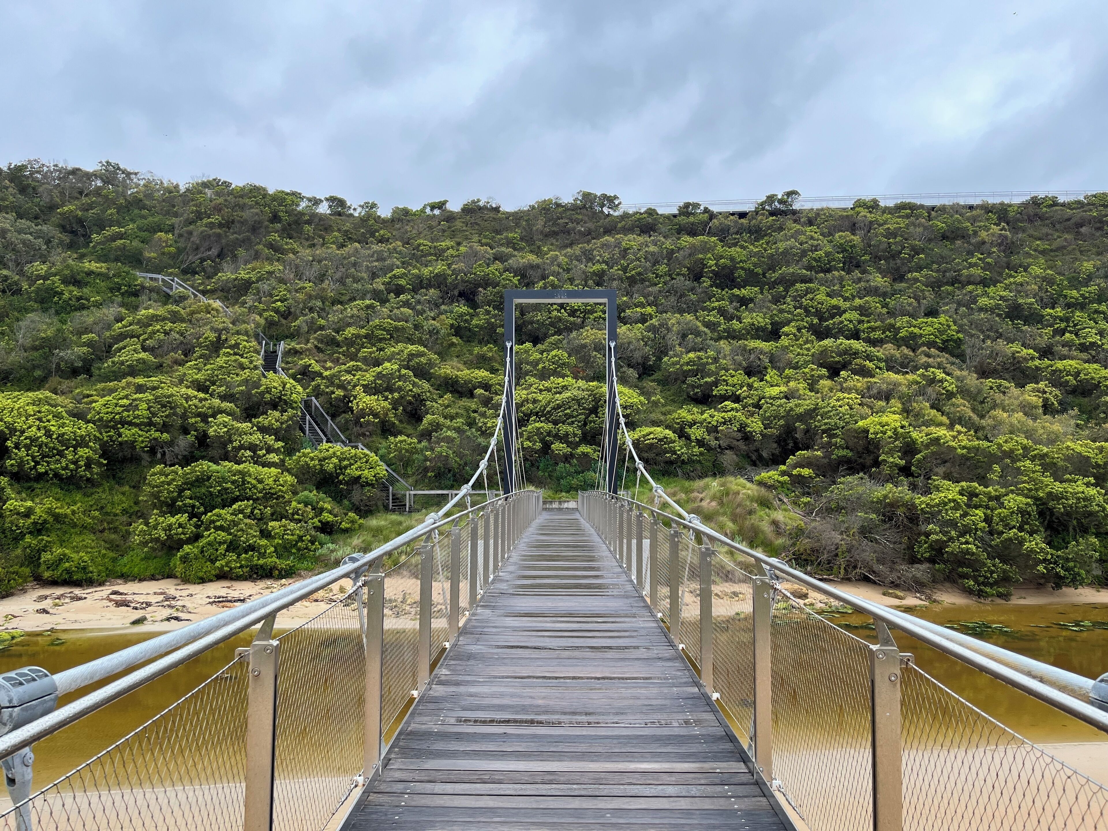 Un puente peatonal de madera con barandillas metálicas, que cruza un río con un promontorio boscoso al fondo.
